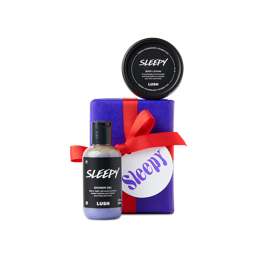 Sleepy. A rectangular gift box wrapped in deep-purple paper and tied with a red ribbon sits vertically. Two lavender-scented products including a shower gel and body lotion are sitting around the box.