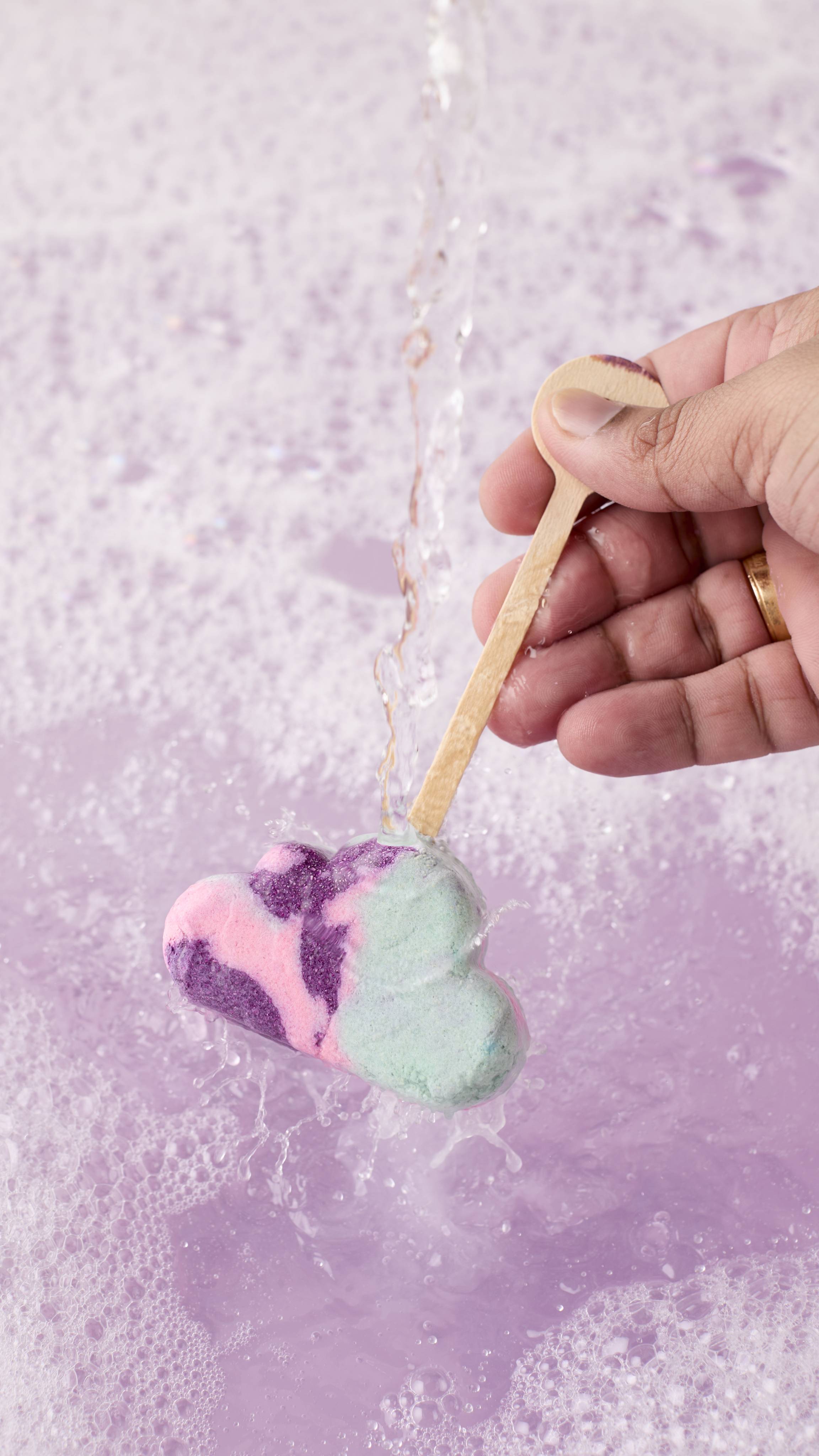 Hand holds a cloud-shaped bubble wand with purple, pink, and mint swirls by its wooden handle. Water pours over it into a purple, bubbly bath.