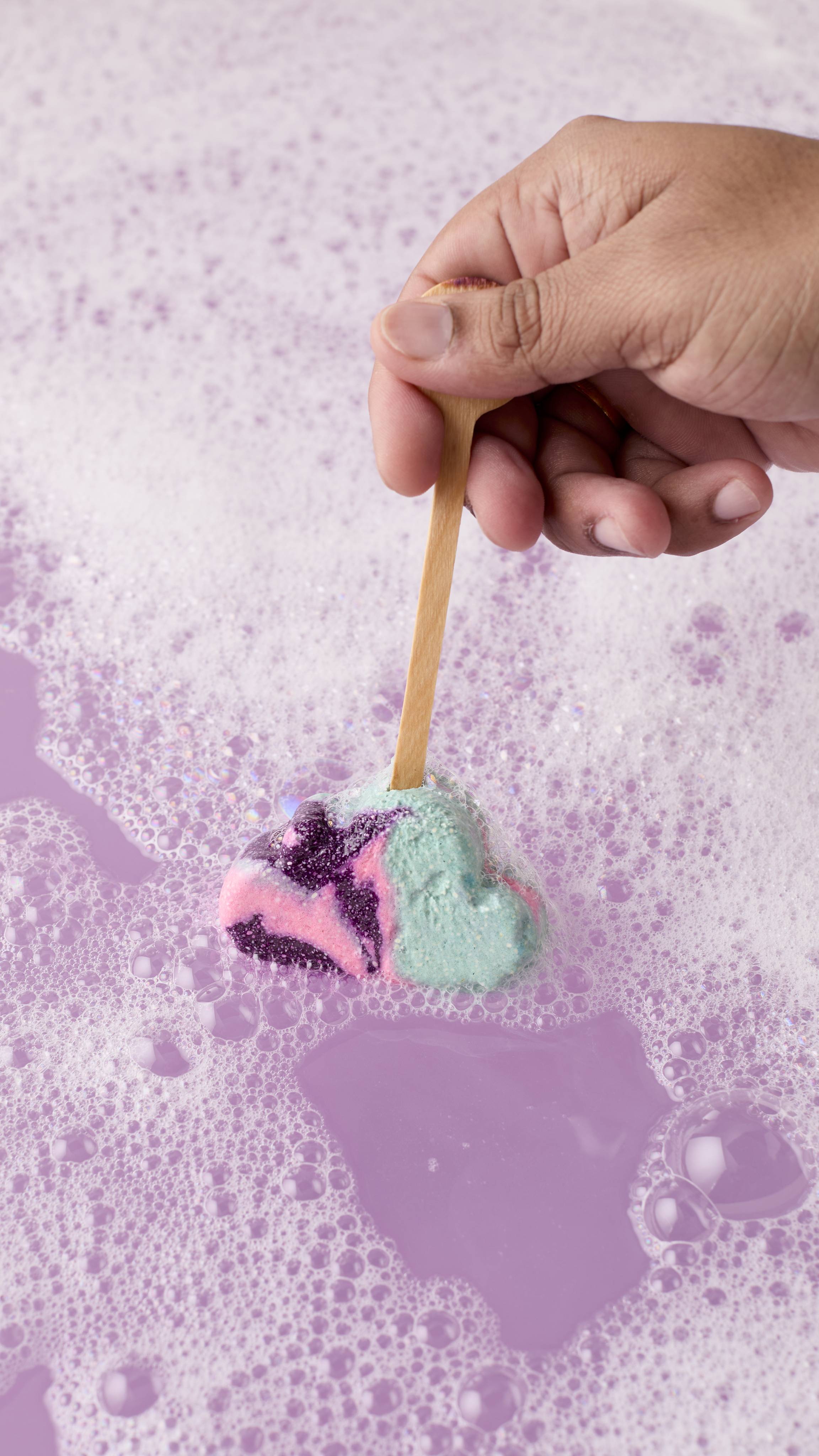 Hand holds a cloud-shaped bubble wand with purple, pink, and mint swirls by its wooden handle. The wand is being swirled into a purple, bubbly bath.