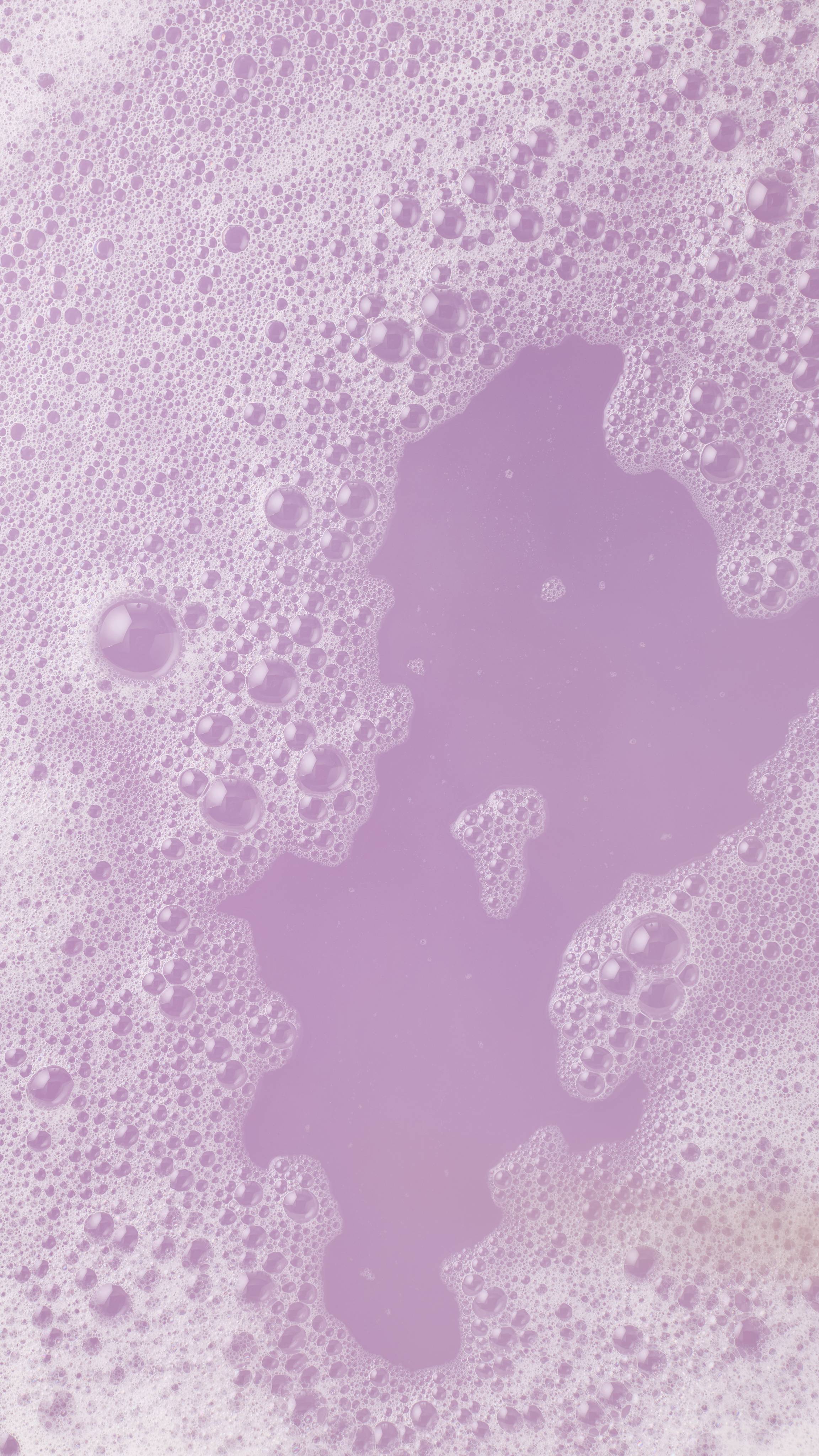 Top-down view of a lavender-colored bath covered in white, frothy bubbles. The soft purple water is visible through a clearing in the center of the foam.