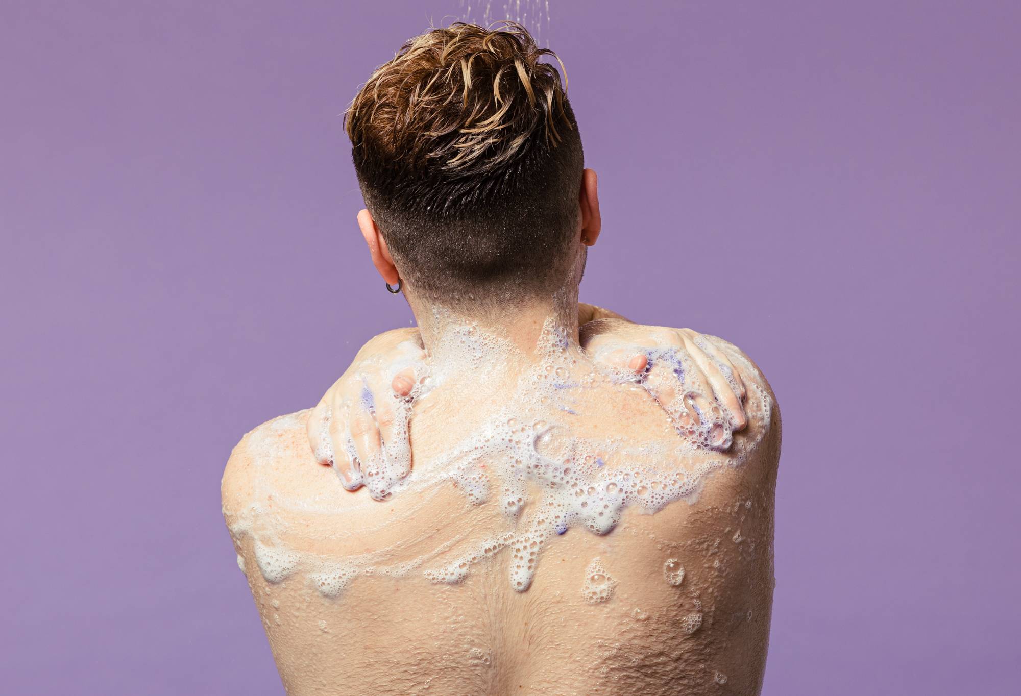 A model stands with upper back and head shown, lather running down from hands over their neck, in front of a purple background.