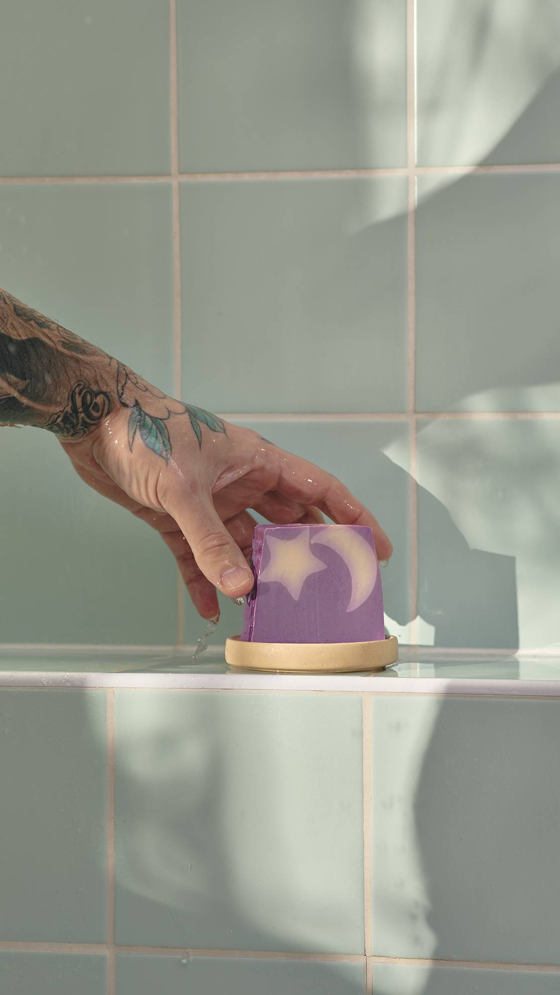A tattooed hand reaches for a purple Sleepy soap bar with a star and moon design. The soap sits in a dish on a light green tiled shower ledge.