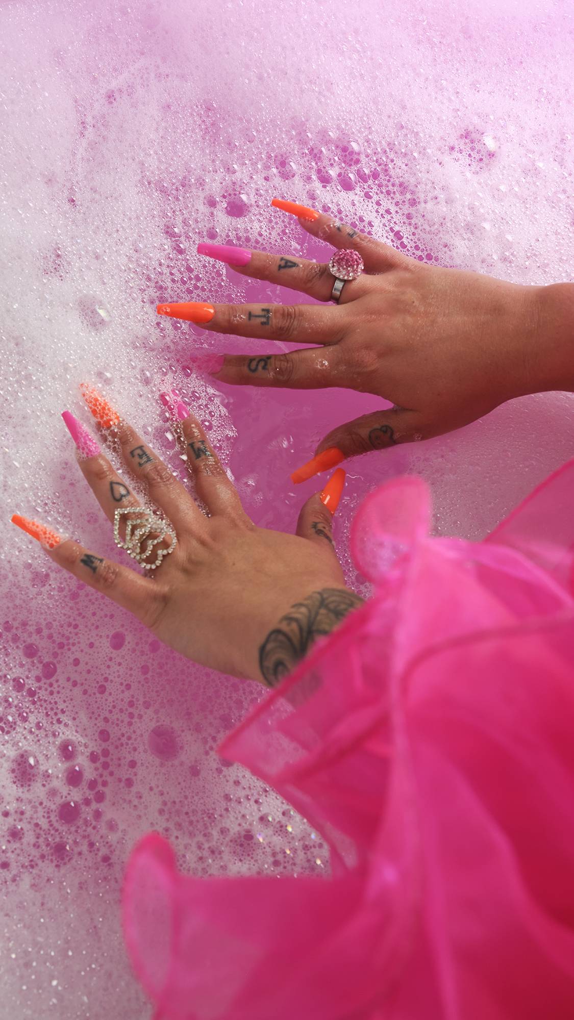 Image shows model’s hands with long orange and pink nails on top of pretty pink waters and bubbles. 