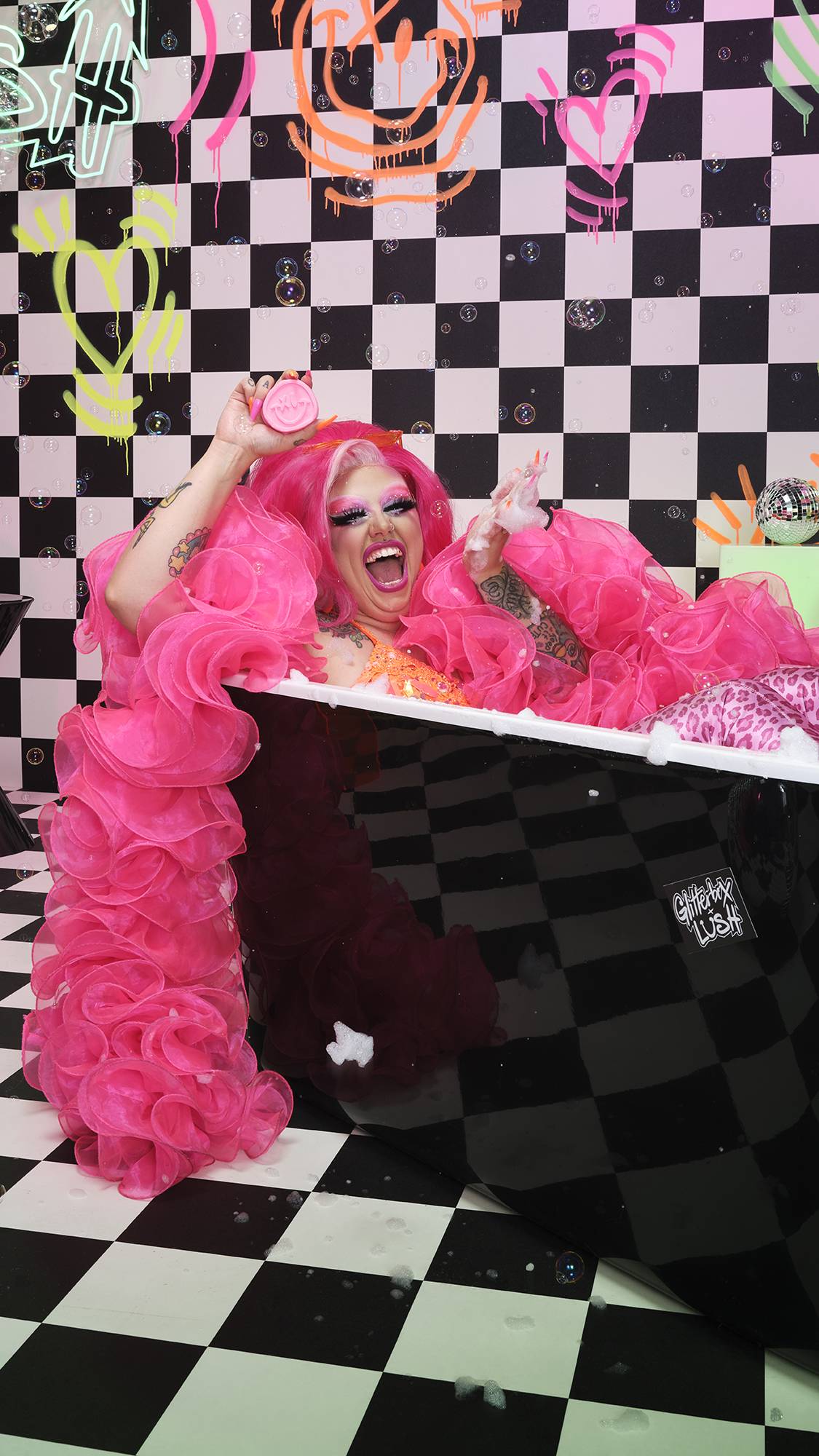 Model is in the bath with a pink ruffled scarf. They are smiling as they hold the bar in front of a checkered and neon wall.