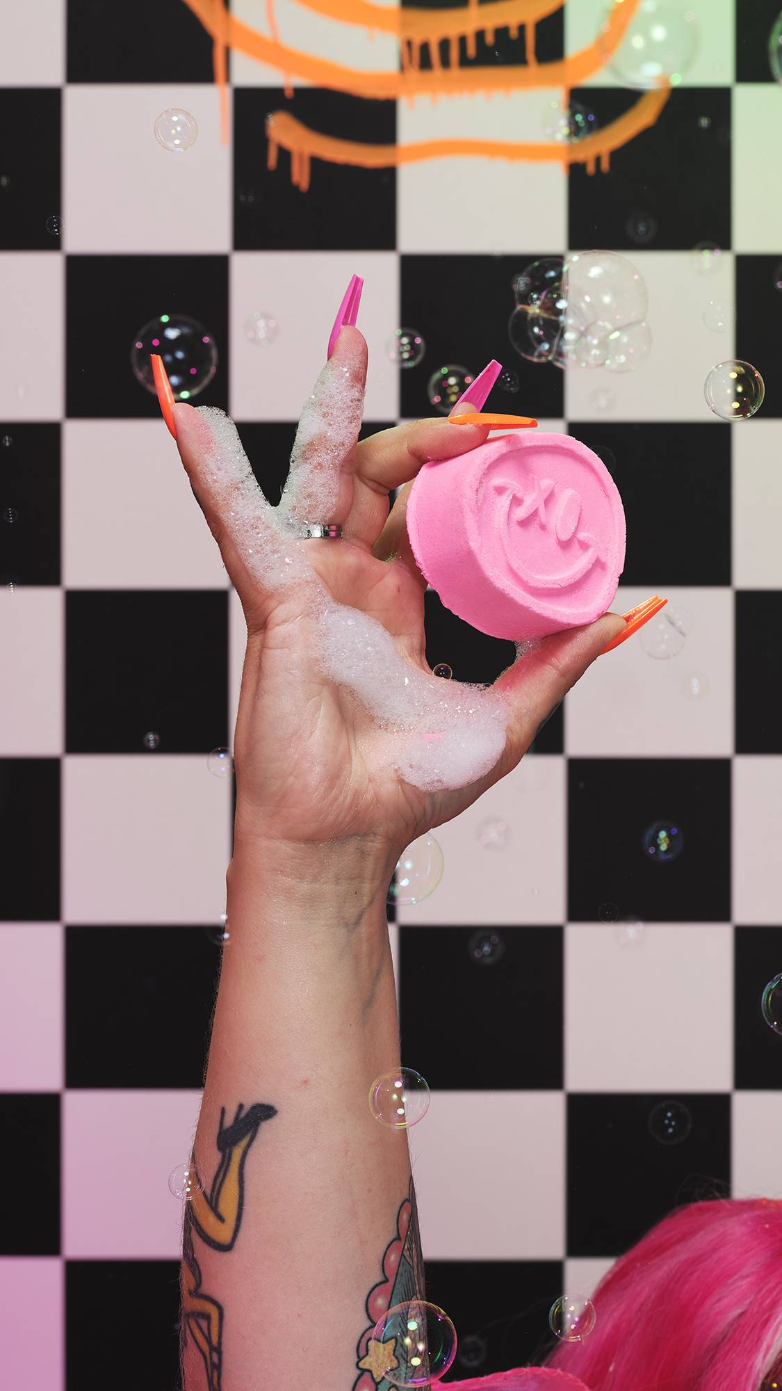 A close-up of the model’s soapy hand holding the pink bubble bar. There is a checkered background and floating bubbles. 
