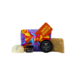 A vibrant fabric-wrapped gift tagged "SMOOTH OPERATOR" sits alongside a scrubee solid body scrub bar, Sticky Dates lip scrub, Happy Skin facial exfoliator, and a bar of soap.