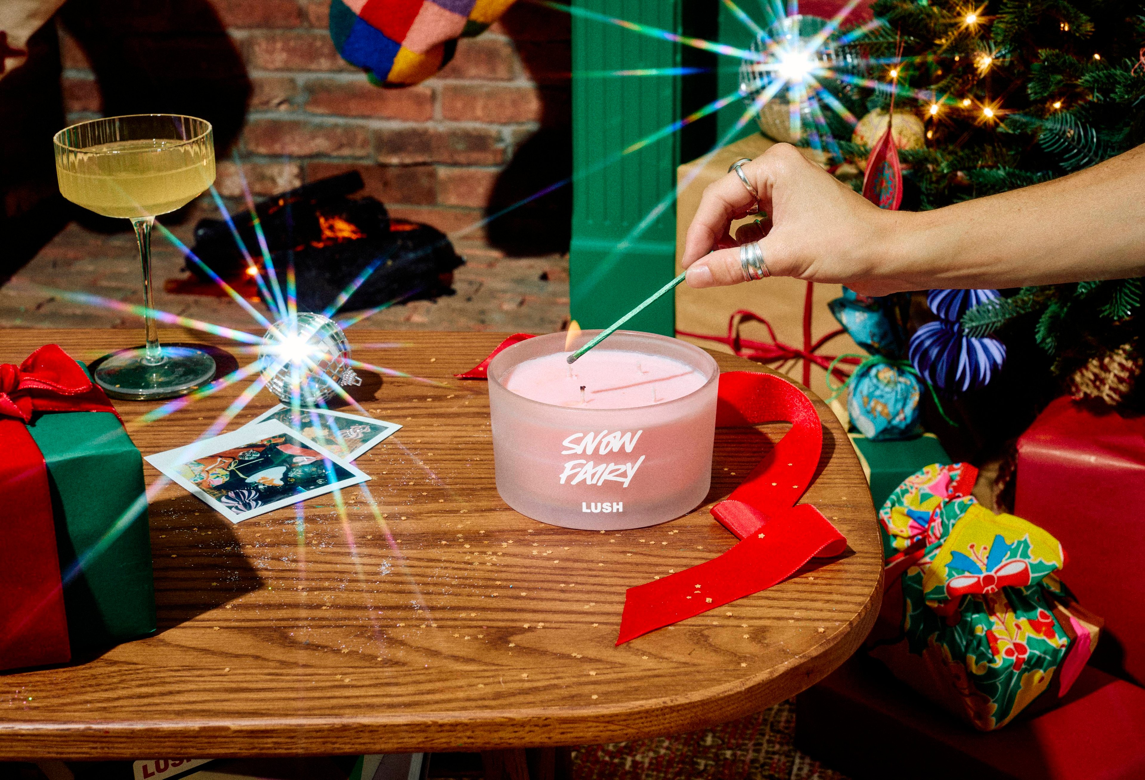 A hand uses a long match to light a large 4-wick Lush Snow Fairy scented candle in a frosted glass jar on a wooden table, styled with red ribbon, a cocktail glass, gifts, and a colourful stocking.