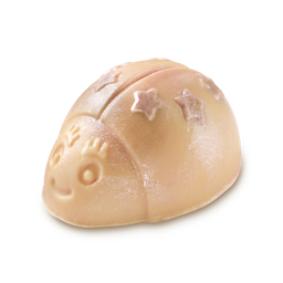 A shiny, light pink LUSH Snow Fairy Scrubee solid body scrub is shown against a light gray background. The product is shaped like a bee with an engraved smiling face and star-shaped indentations on its back, all covered in a fine glitter.