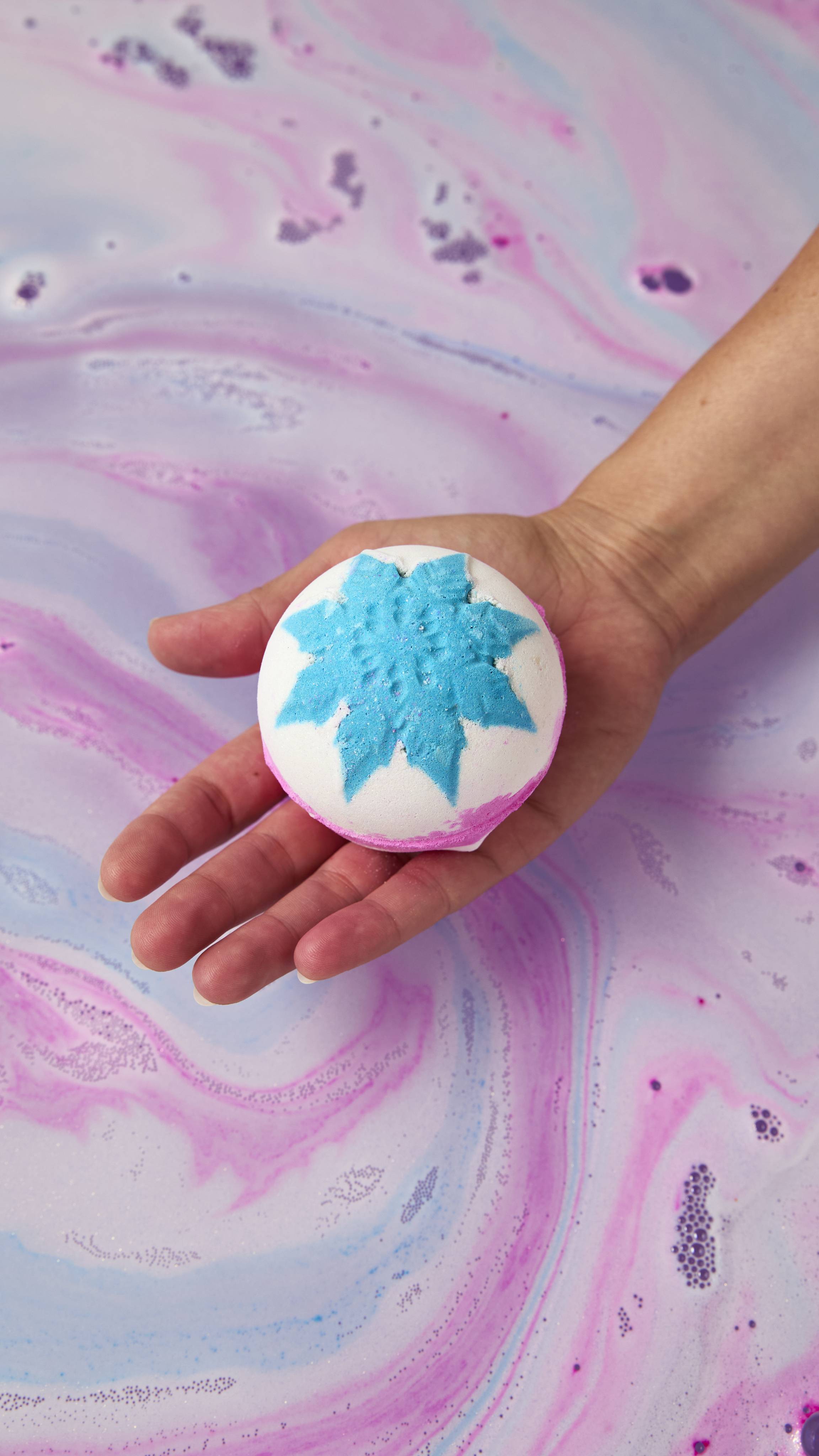 A hand holds a round white "Snow Fairy" bath bomb decorated with a bright blue snowflake pattern on top and a pink base, held over swirling pastel pink, blue, and purple bathwater.