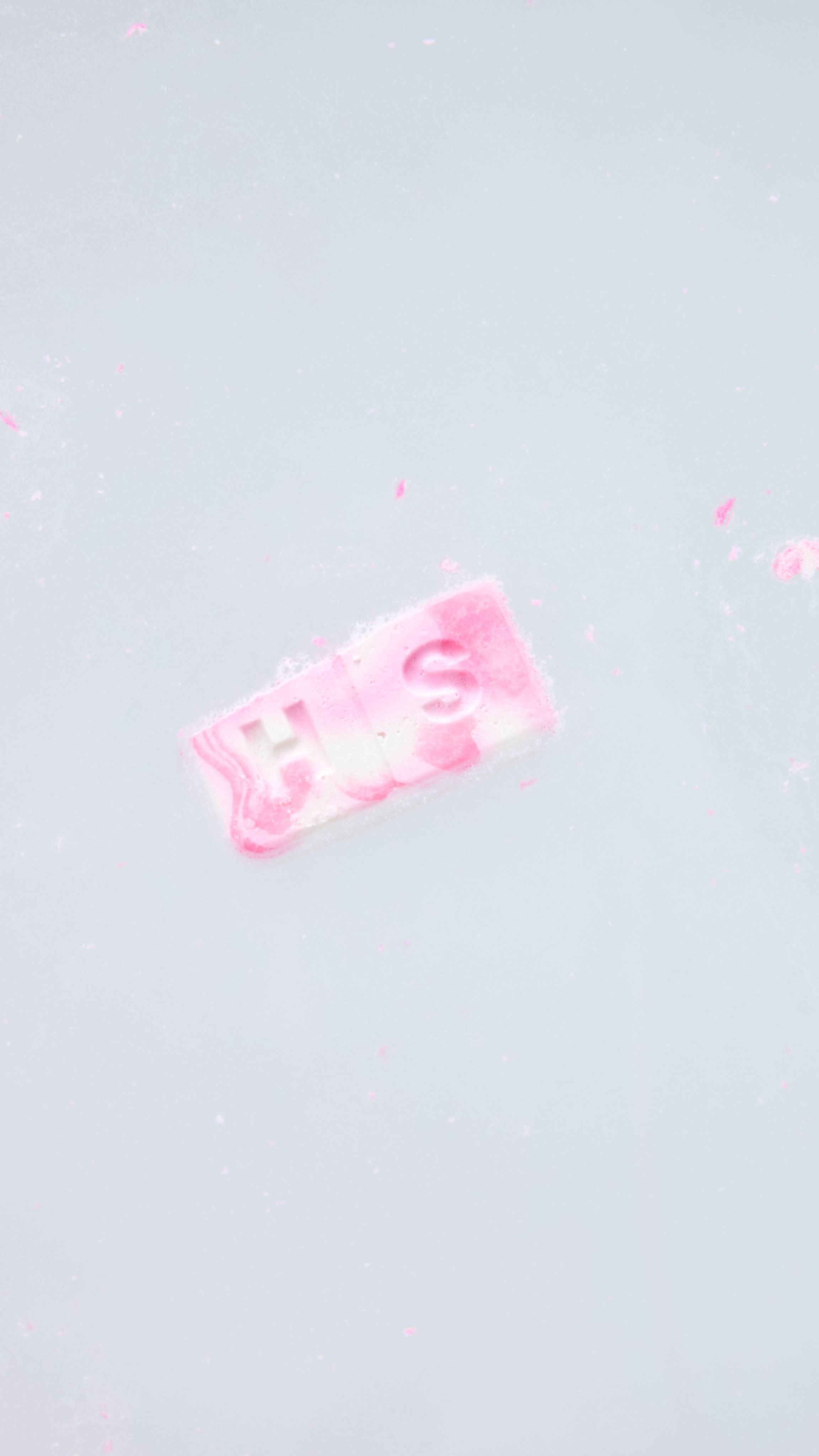 A piece of a pink and white marbled wax melt embossed with the letters "HS" dissolves into warm water, surrounded by faint swirls of pink colour and soft foam.