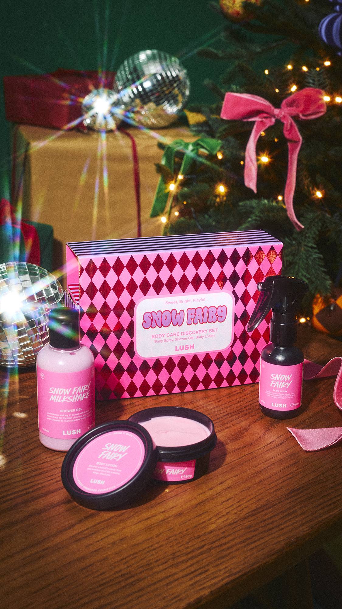An open Lush 'Snow Fairy Body Care Discovery Set' on a wooden surface, surrounded by its contents: shower gel, body spray, and an open lotion tub, set against disco balls and a decorated Christmas tree.  