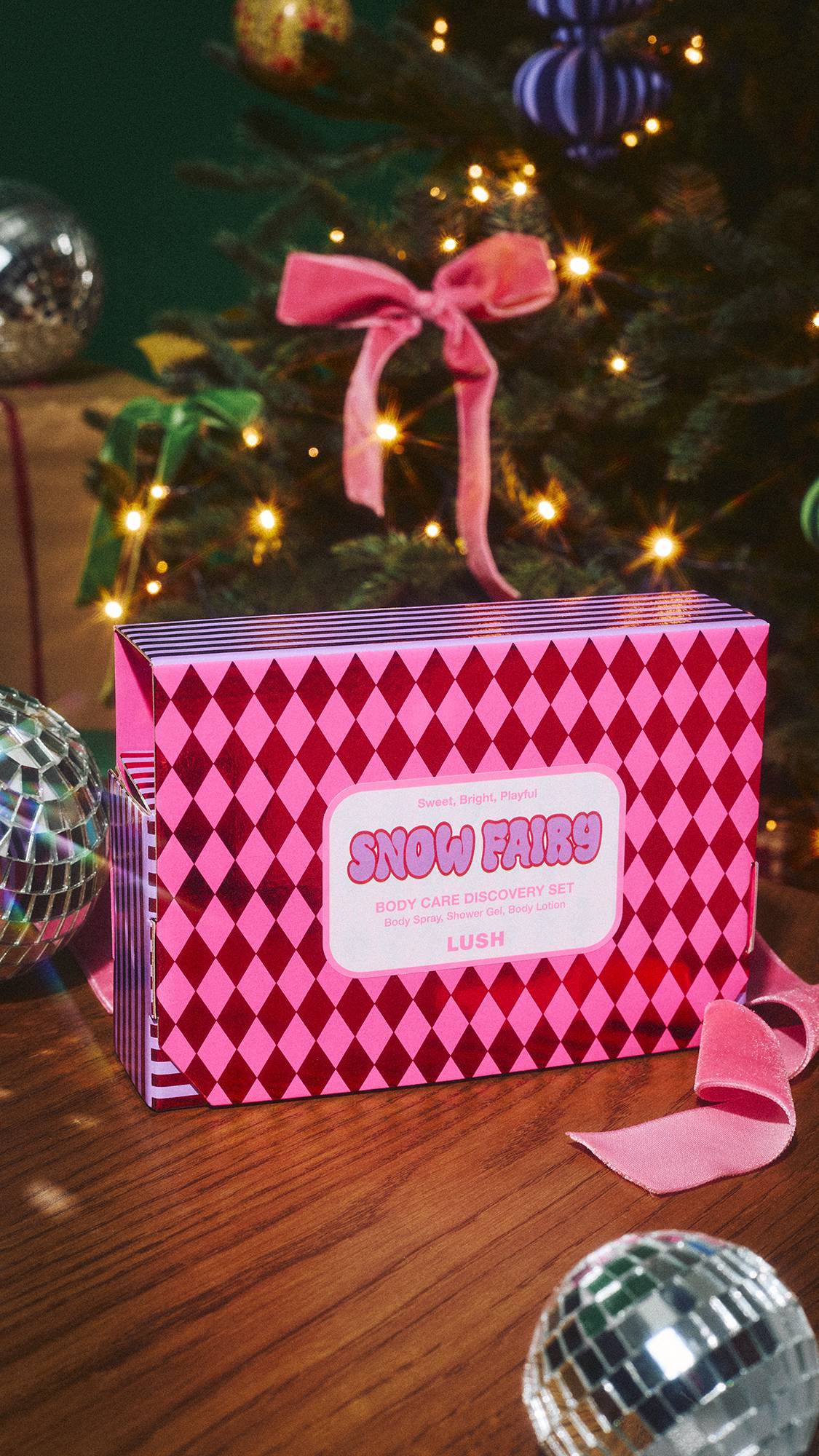 A Lush 'Snow Fairy Body Care Discovery Set' gift box, featuring a pink diamond pattern, sits on a wooden surface with a pink velvet ribbon and disco balls, set in front of a twinkling Christmas tree.  