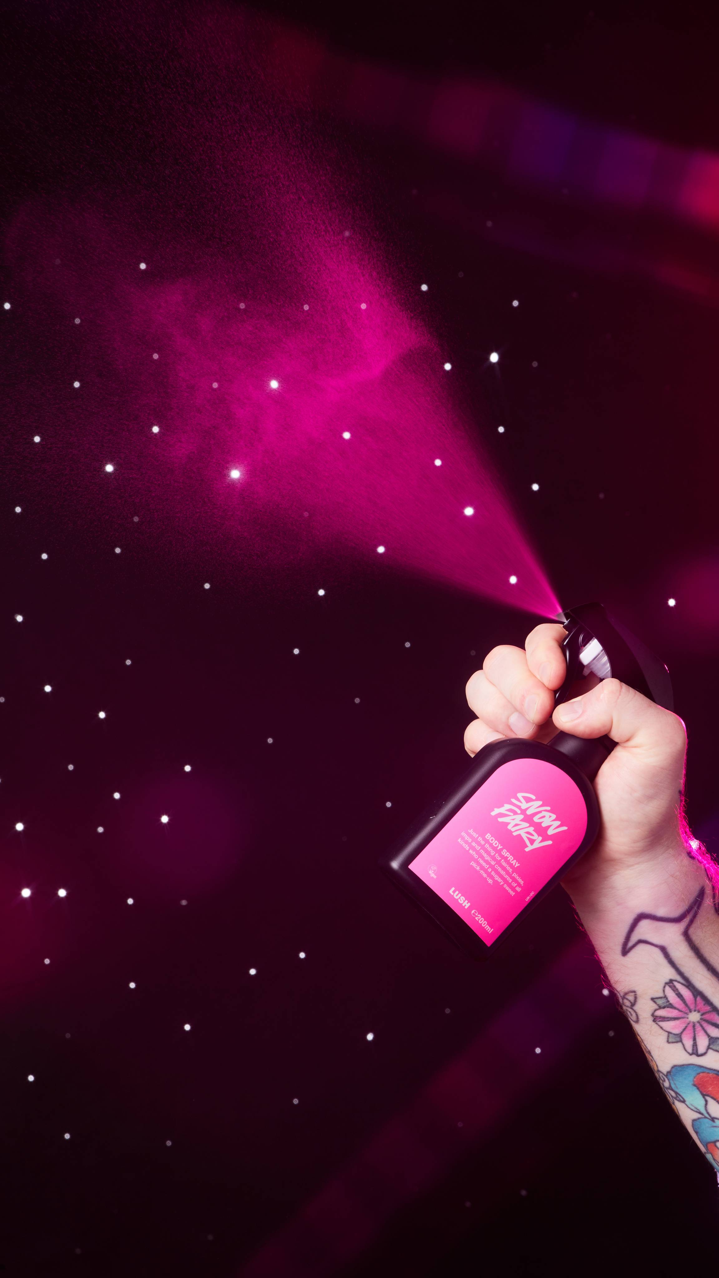 A close-up of the model's hand spritzing the Snow Fairy body spray in the air on a starry, pink-lit background. 