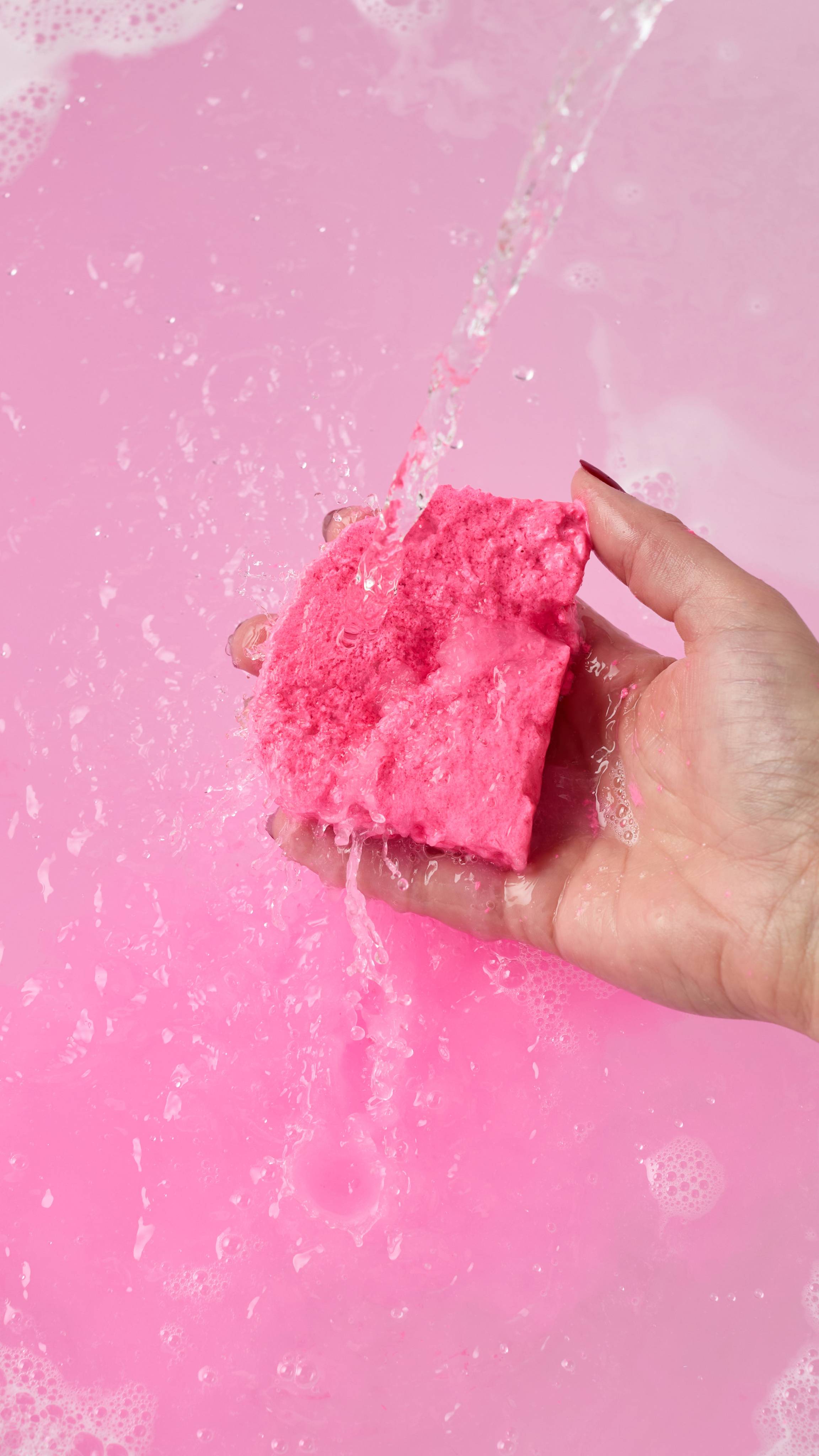 The image shows the model's hand reaching in from the right as it holds the Snow Fairy Bubble Mountain bubble bar in front of running tap water. The water has been turned a pastel pink colour from the product and is scattered with bubbles.