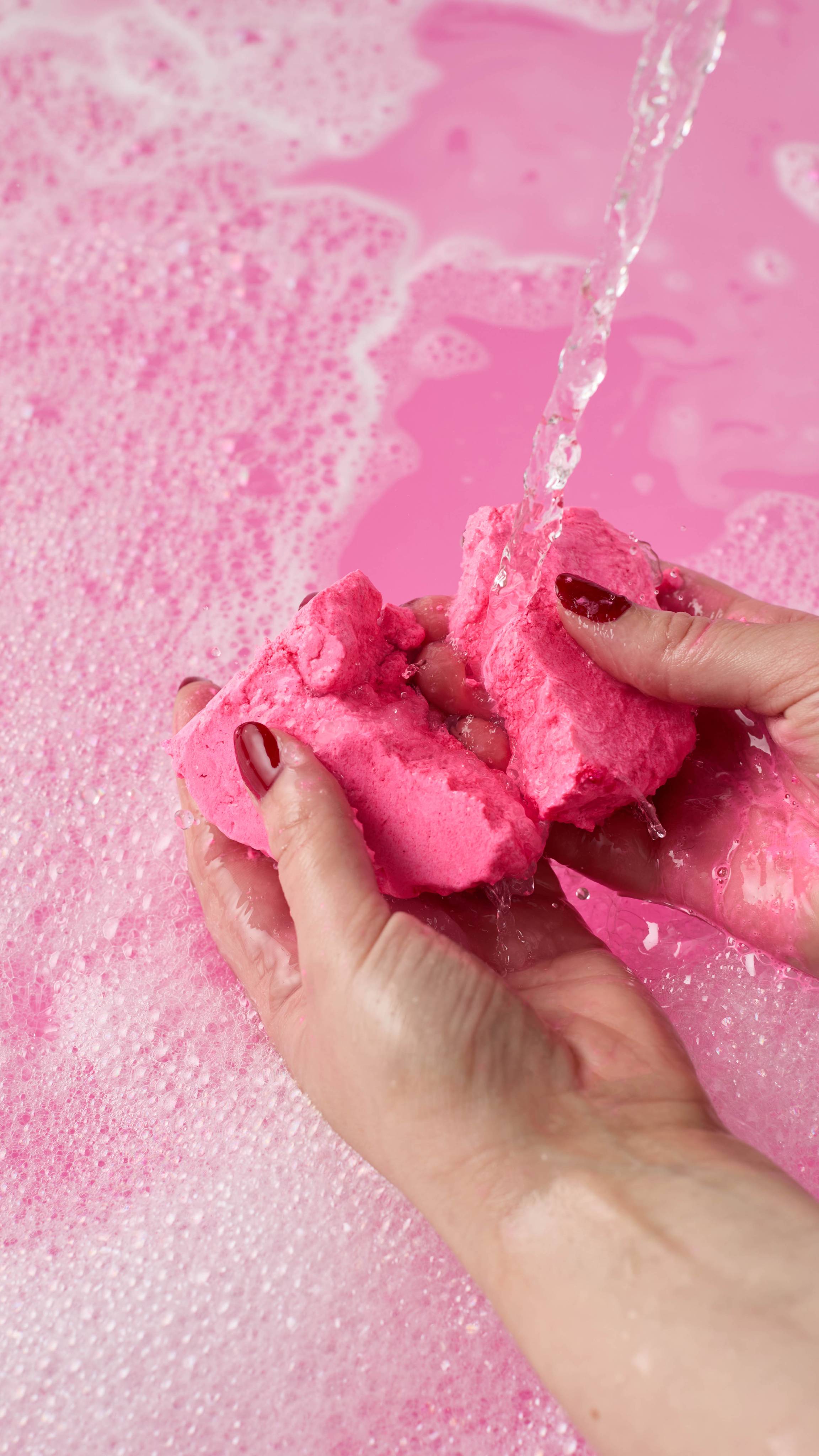 The image shows the model using both hands to break the Snow Fairy Bubble Mountain bubble bar in half underneath running tap water. The water has been turned a bright pink colour from the product with a layer of thick bubbles.