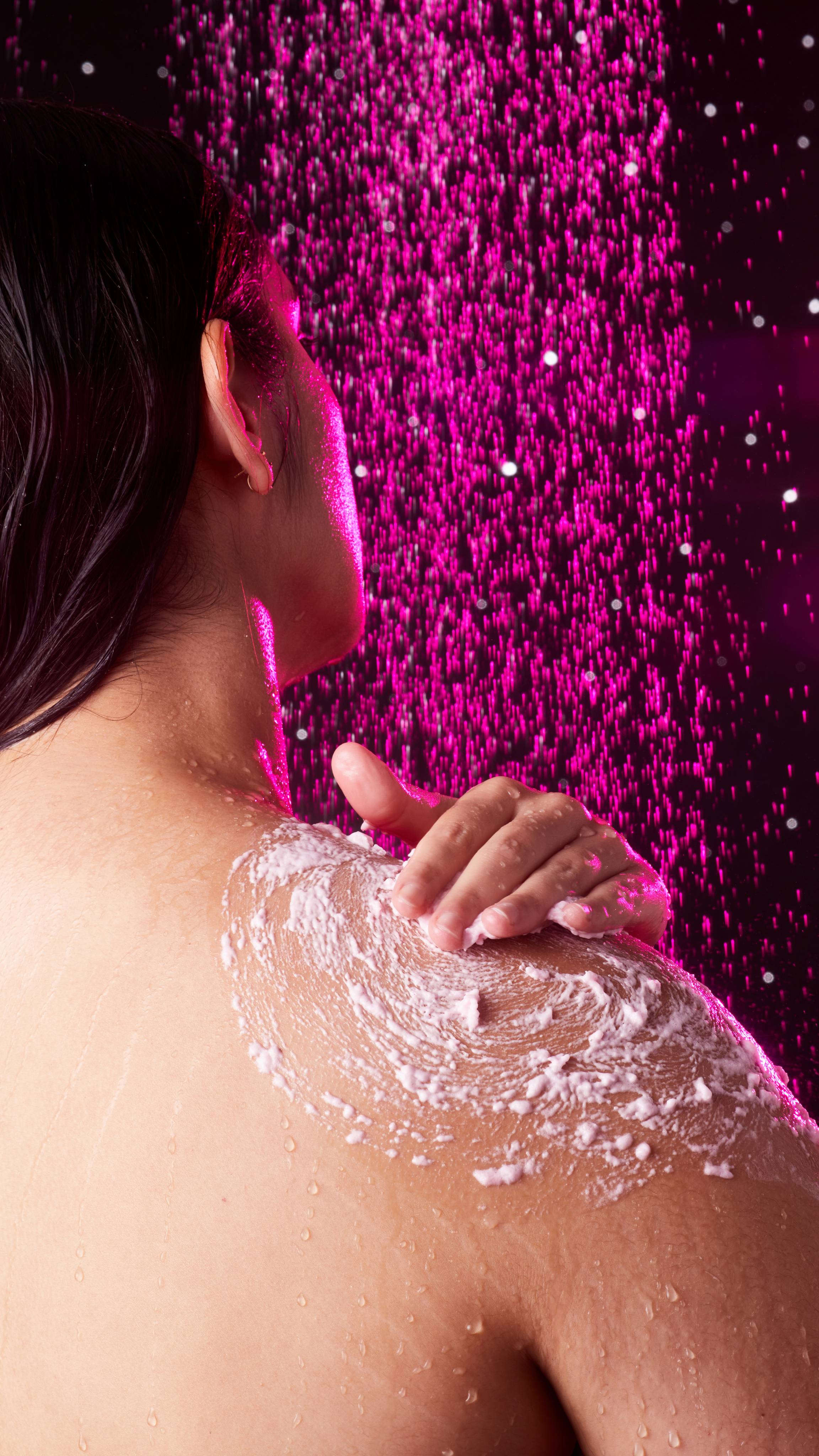 Model is in the shower facing away as they reach to exfoliate their shoulder with the product in circular motions.