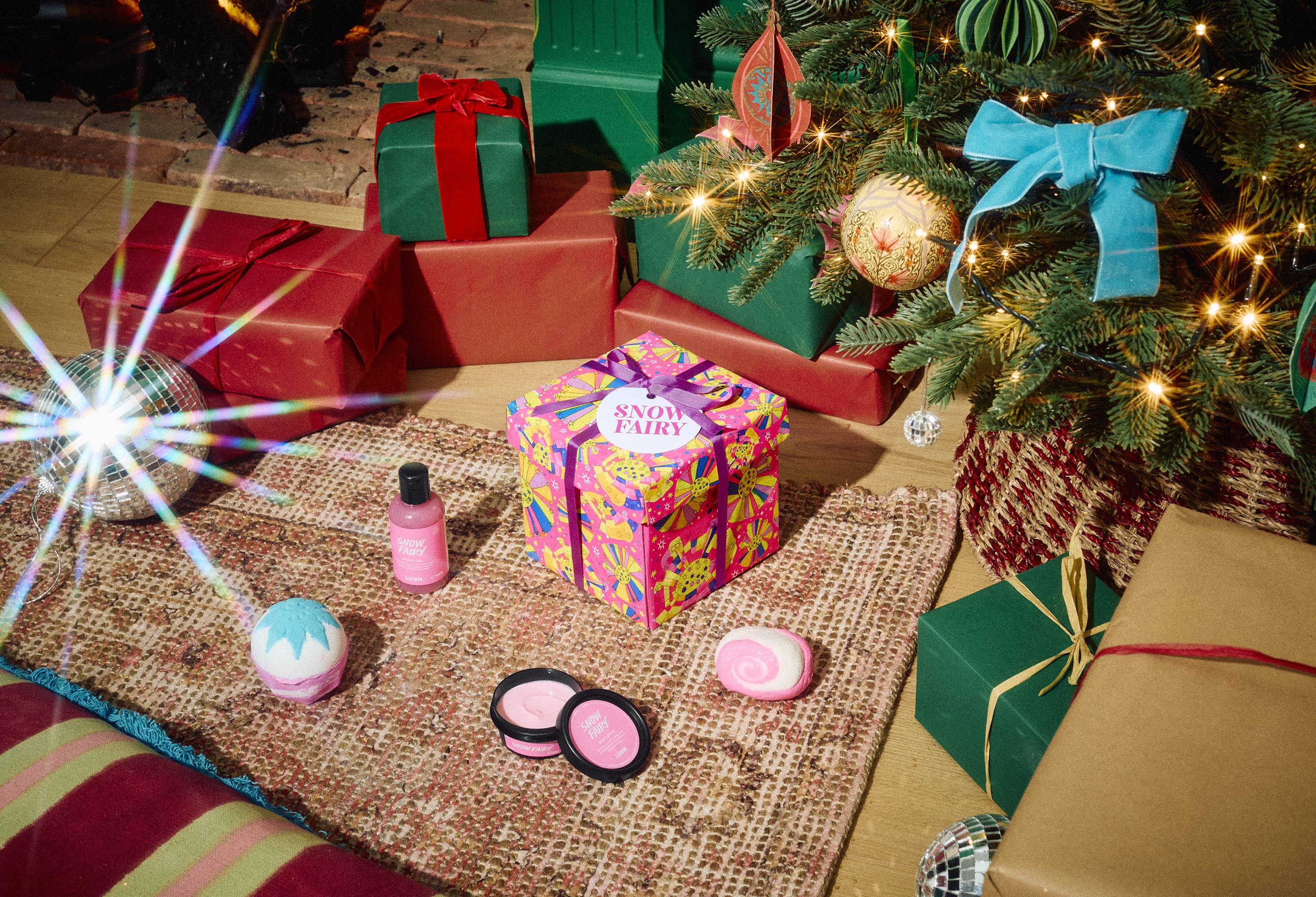 The Lush Snow Fairy gift box sits on a patterned rug surrounded by pink shower gel, body lotion, a bath bomb, and a bubble bar, set in a festive room with wrapped presents and a decorated Christmas tree.
