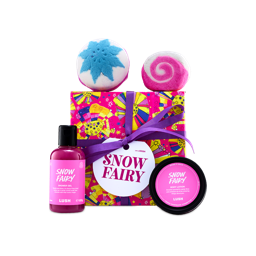 A Lush "Snow Fairy" gift set wrapped in bright patterned paper with a purple ribbon, surrounded by pink shower gel, body lotion, a blue snowflake bath bomb, and a pink swirl bubble bar.
