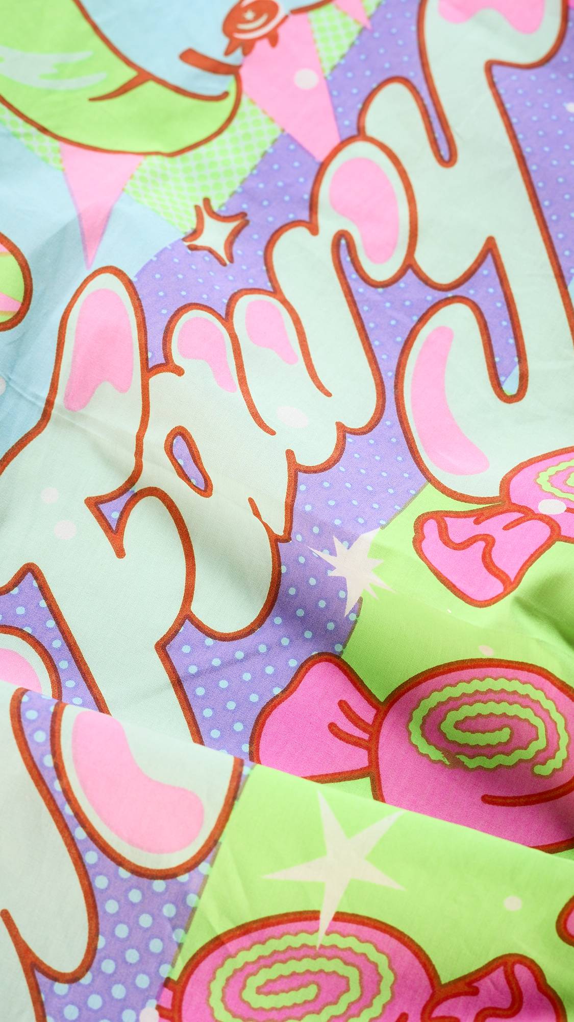 A close-up of the "Snow Fairy" Knot Wrap, a reusable fabric from Lush. The pattern features a whimsical, colorful collage with "Fairy" written in a bubbly font and designs of candies, stars, and checkered patterns.