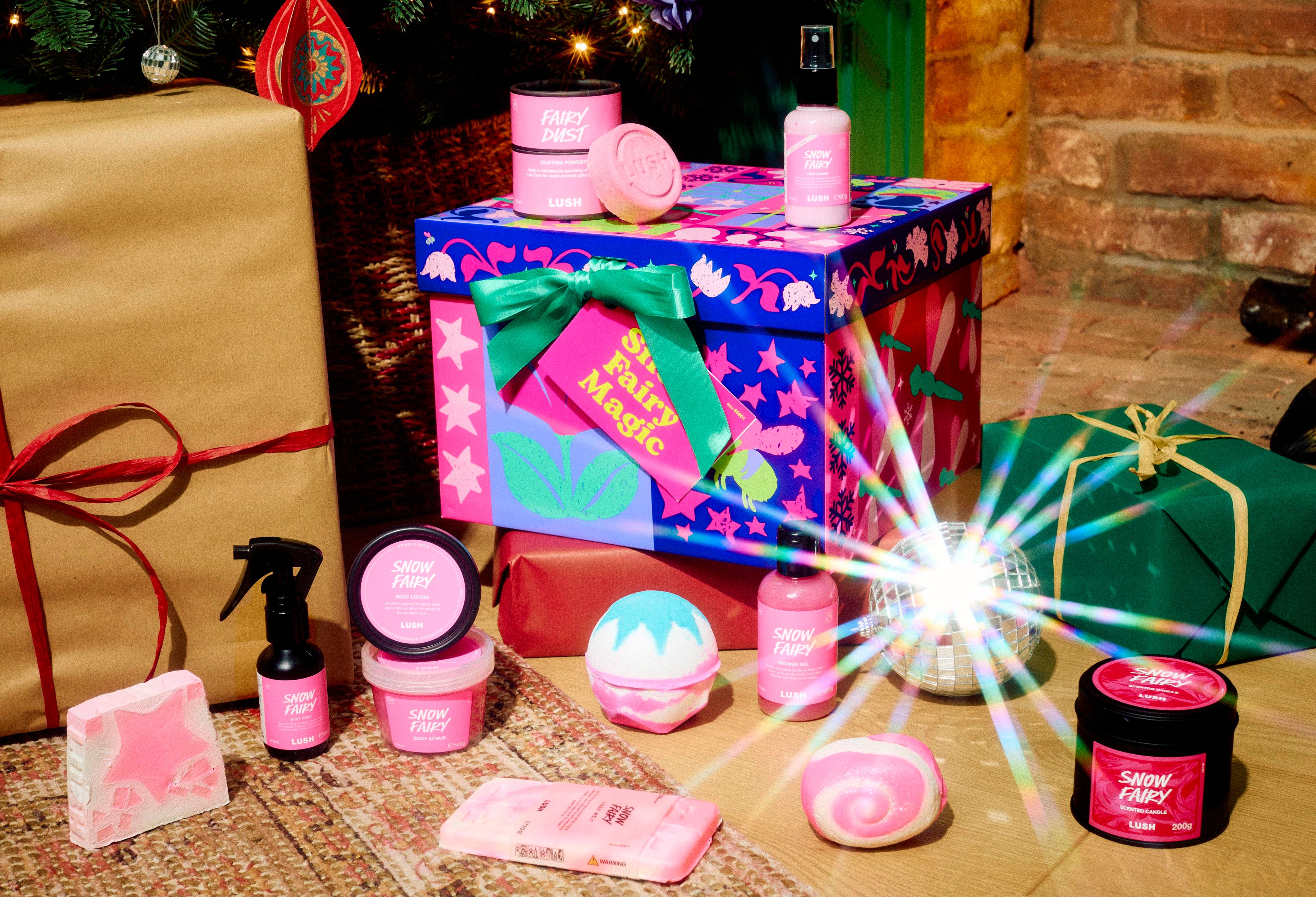 The Lush Snow Fairy Magic gift box rests on a patterned rug surrounded by twelve Snow Fairy products, including shower gel, candle, body lotion, dusting powder, and bath items, in a cosy room setting near wrapped presents.