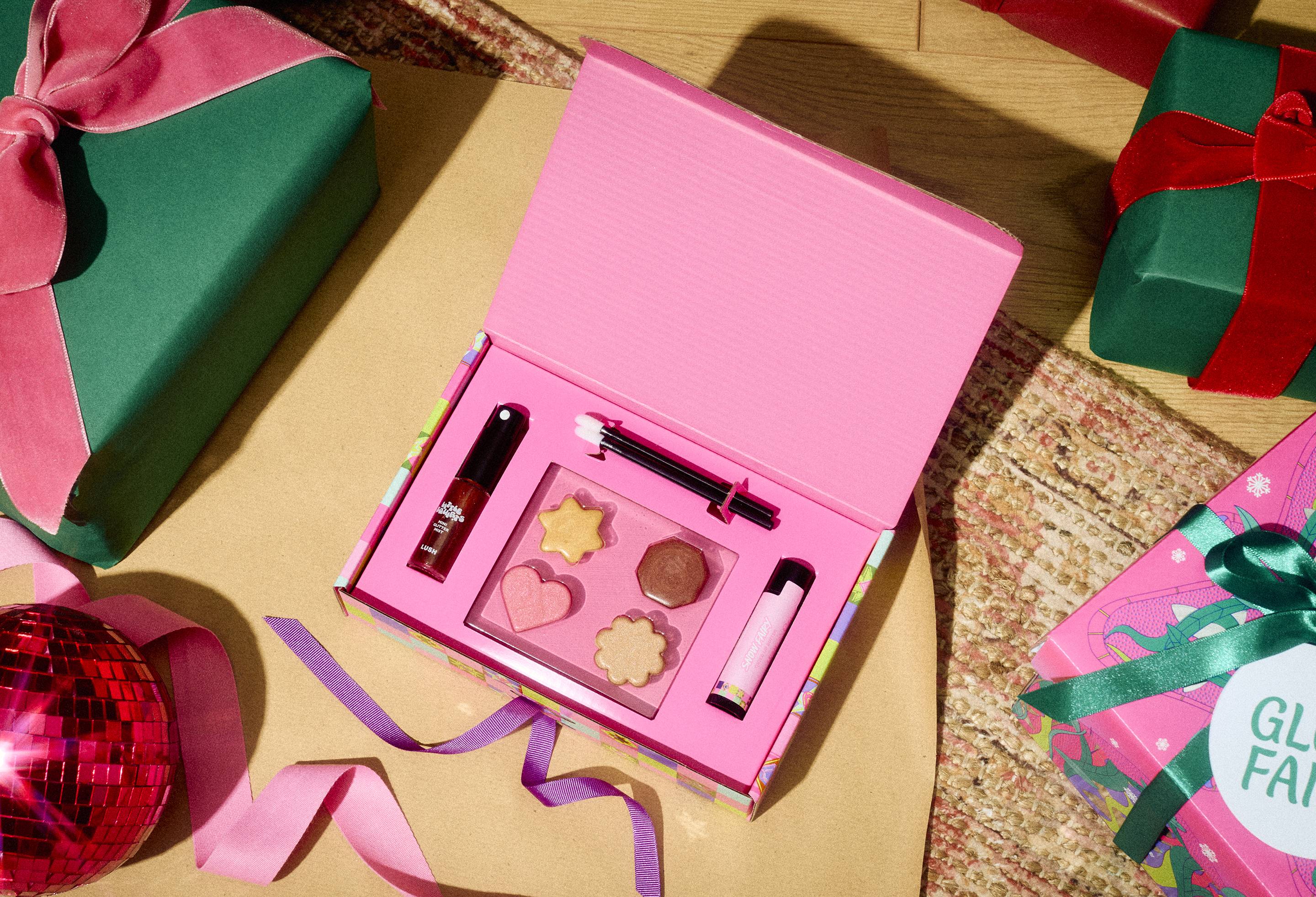 An open Lush Snow Fairy Make Up Set box displaying shimmer cosmetics, lip butter, and glitter spray, set on a floor surrounded by wrapped presents, ribbons, and a pink bauble in a festive room setting.