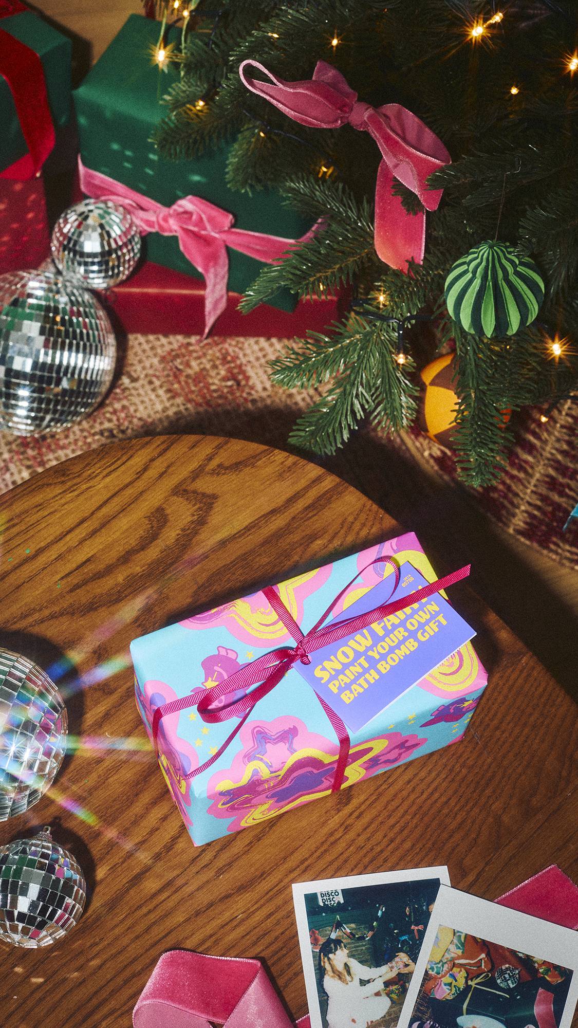 A Lush 'Snow Fairy Paint Your Own Bath Bomb Gift' box, wrapped in vibrant pink and blue paper tied with a pink ribbon, sits on a wooden table beside disco balls beneath a decorated Christmas tree.