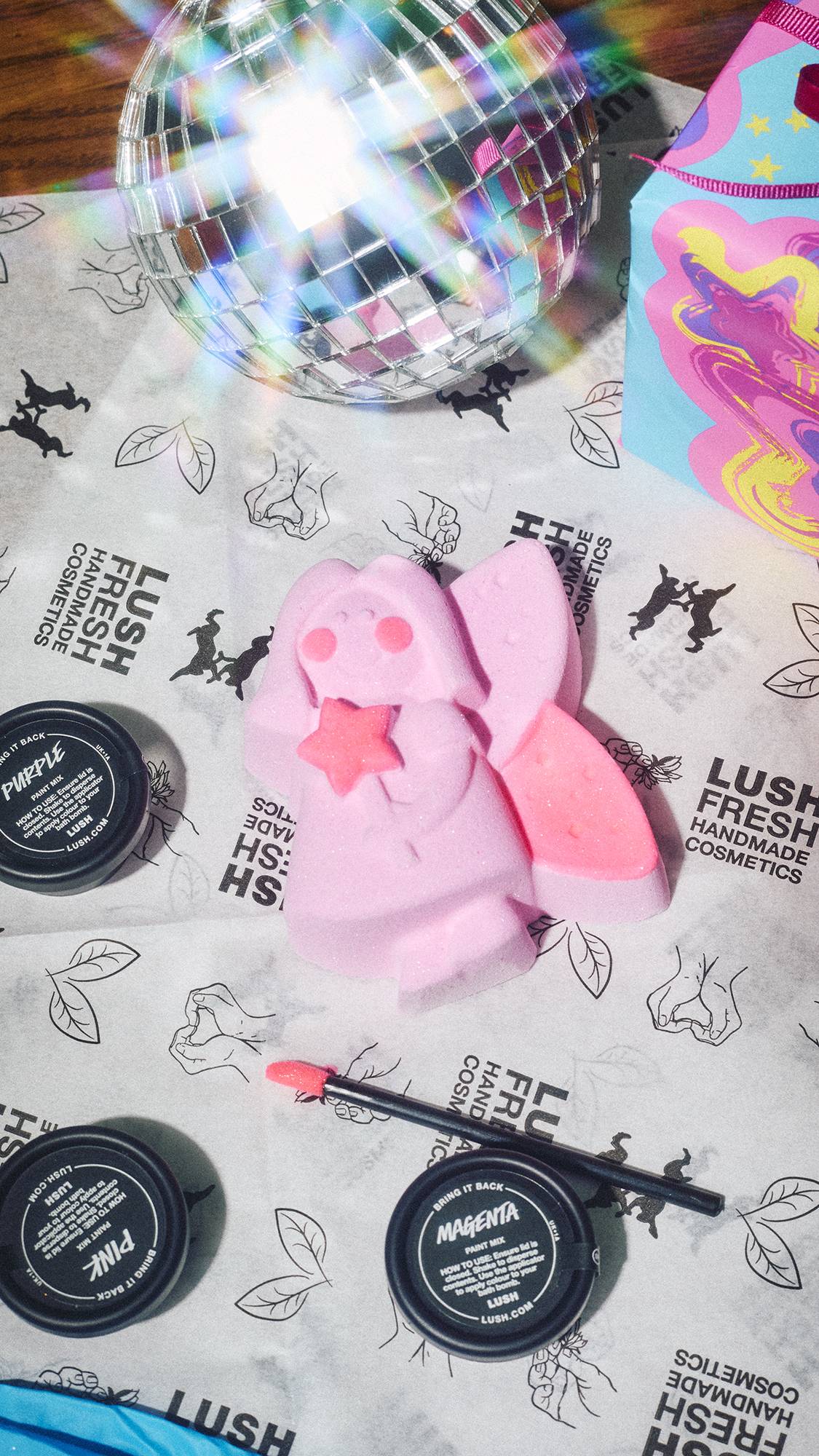 A hand-painted Lush Snow Fairy bath bomb with pink accents lies on branded tissue paper alongside three colour paint pots, a brush, and a glittering disco ball.  