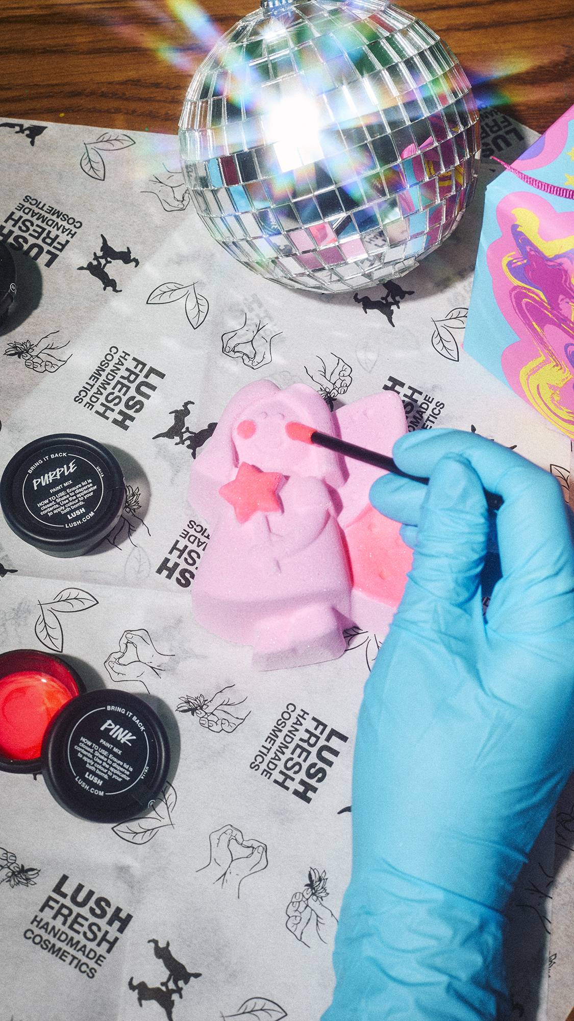 A gloved hand uses a brush to apply pink paint to a Lush Snow Fairy bath bomb, surrounded by paint pots, Lush wrapping paper, and a glittering disco ball.  