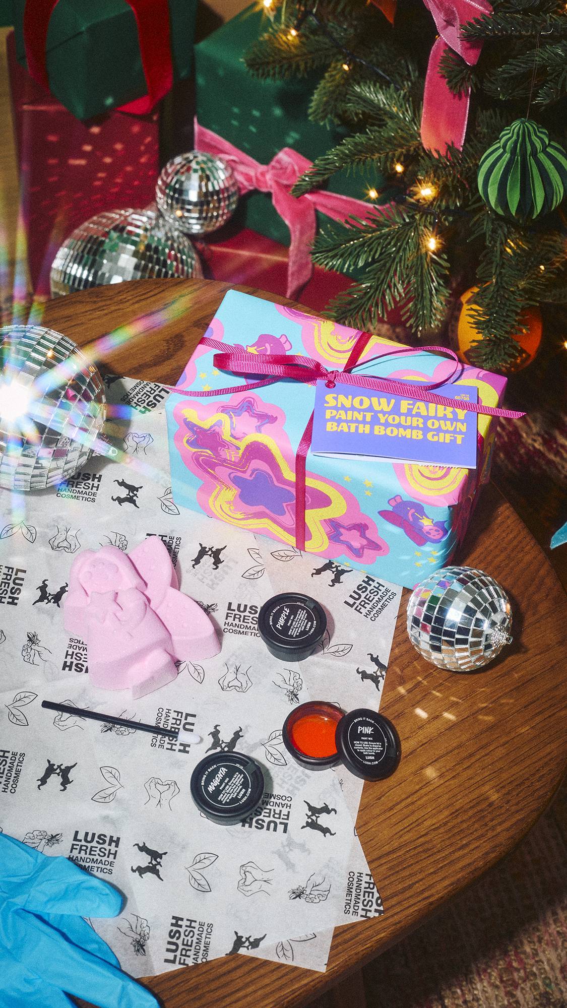 An unwrapped Lush 'Snow Fairy Paint Your Own Bath Bomb Gift' set on a wooden table, featuring a pink fairy bath bomb, paint pots, brush, and wrapped box beside a mini disco ball under a Christmas tree.  