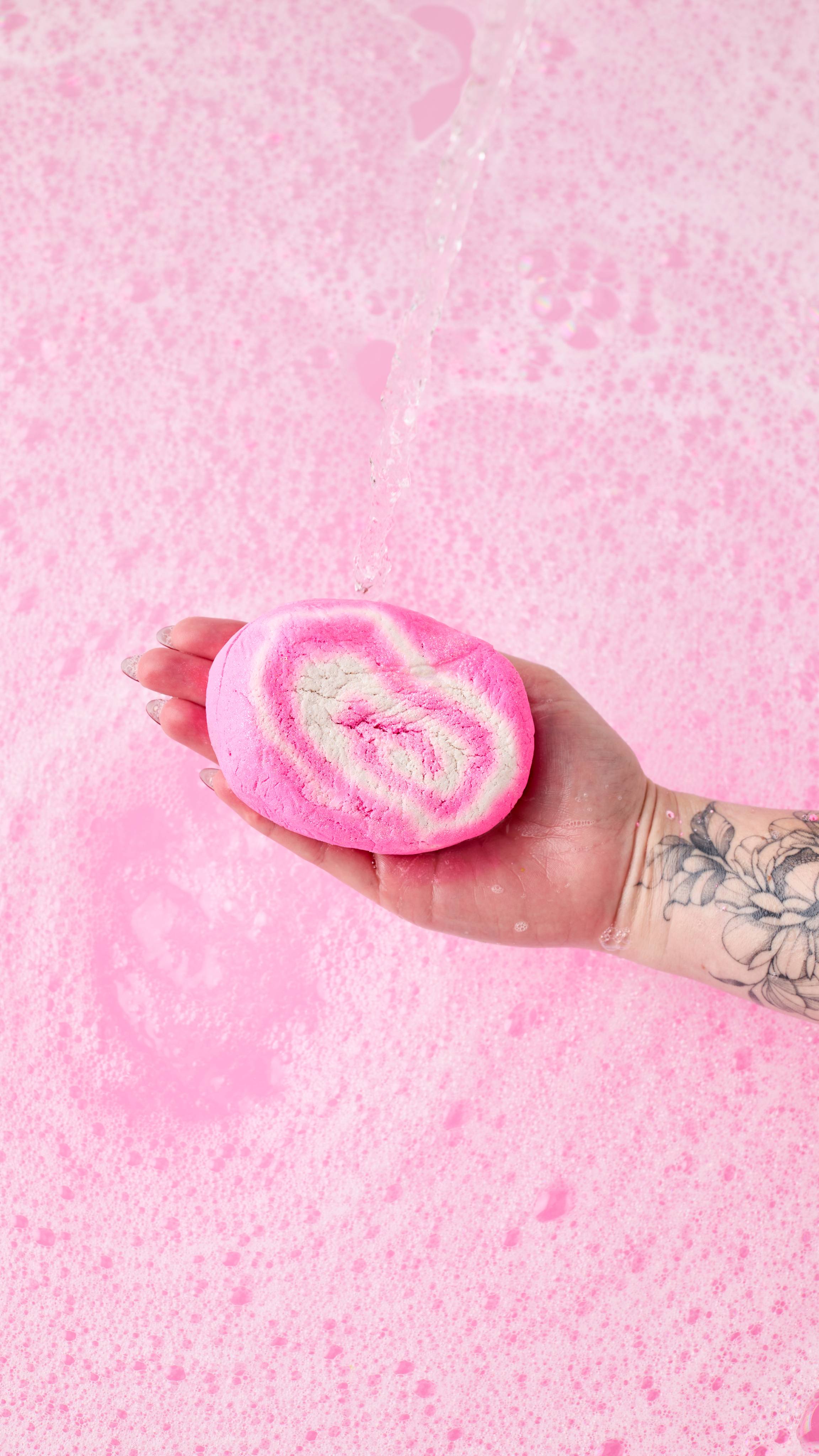 A hand with a tattooed forearm holds a Snow Fairy Roll bubble bar under a stream of water. The pink and white bubble bar is held over a frothy, pink-tinted bath.
