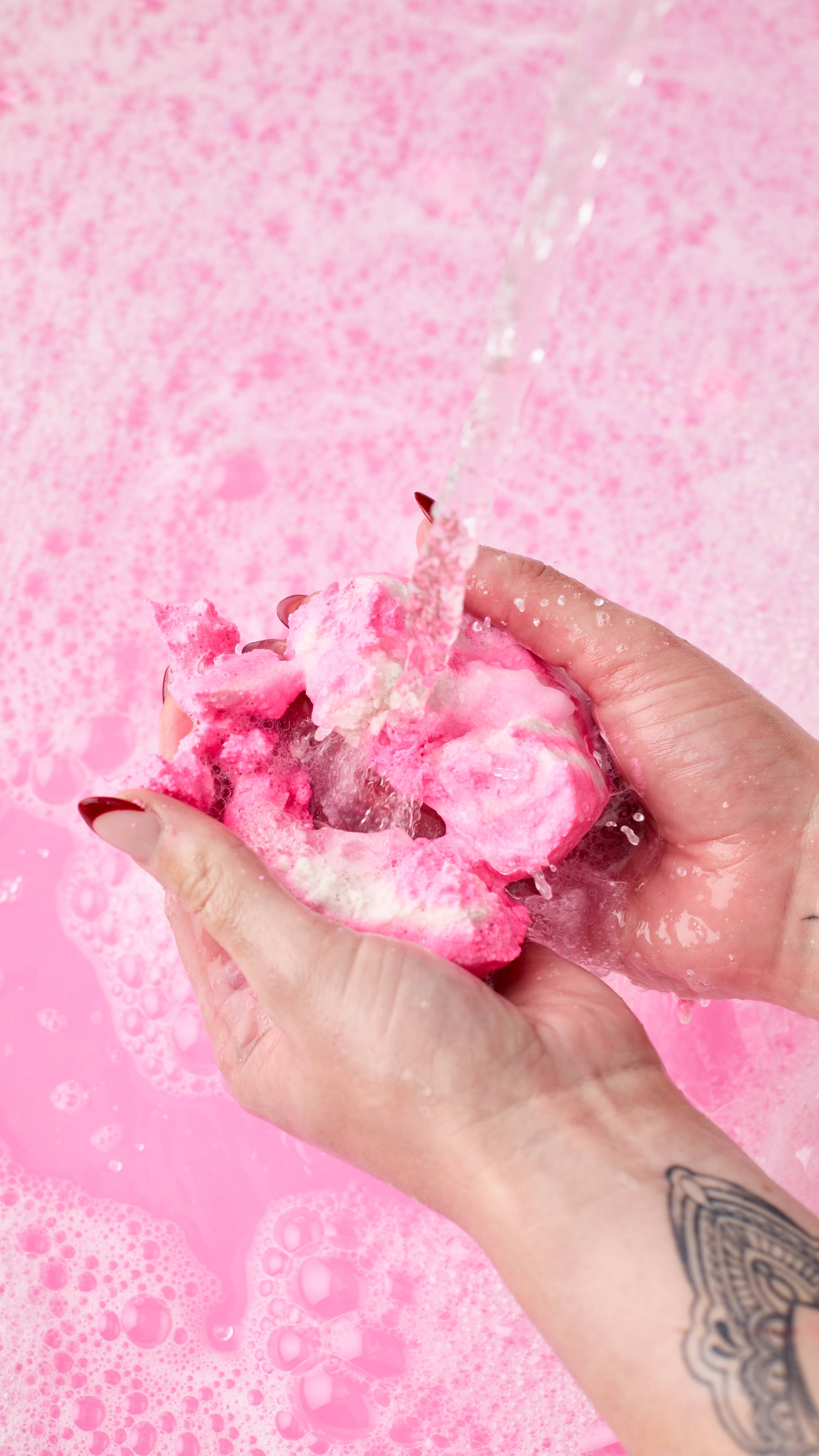 The image shows light-toned hands gently crumbling a piece of The Big Snow Fairy Roll bubble bar under a stream of water. The pink and white bubble bar is held over a frothy, pink-tinted bath.
