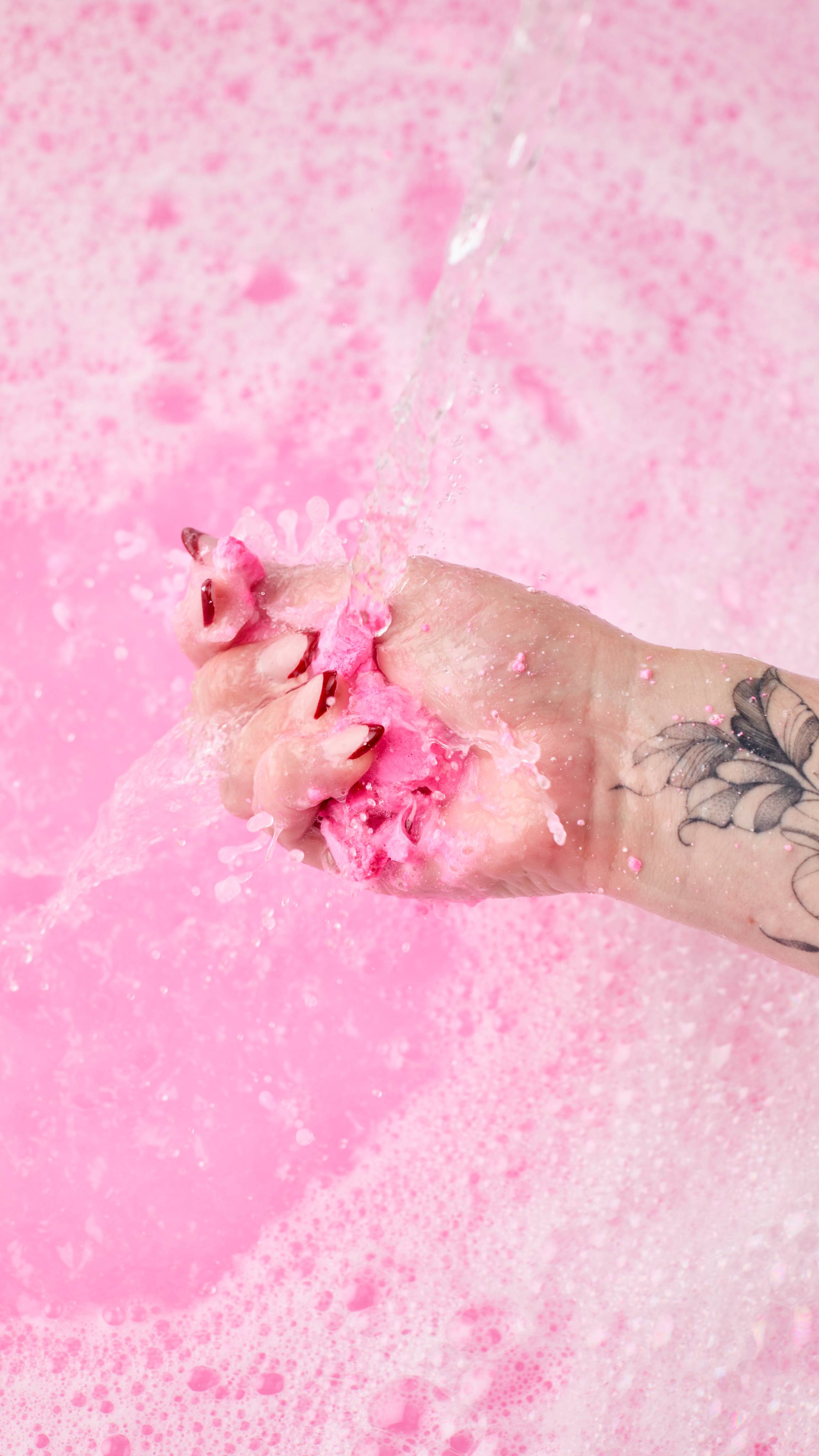The image shows a light-toned hand gently crumbling the Snow Fairy Roll bubble bar under a stream of water. The pink and white bubble bar is held over a frothy, pink-tinted bath.