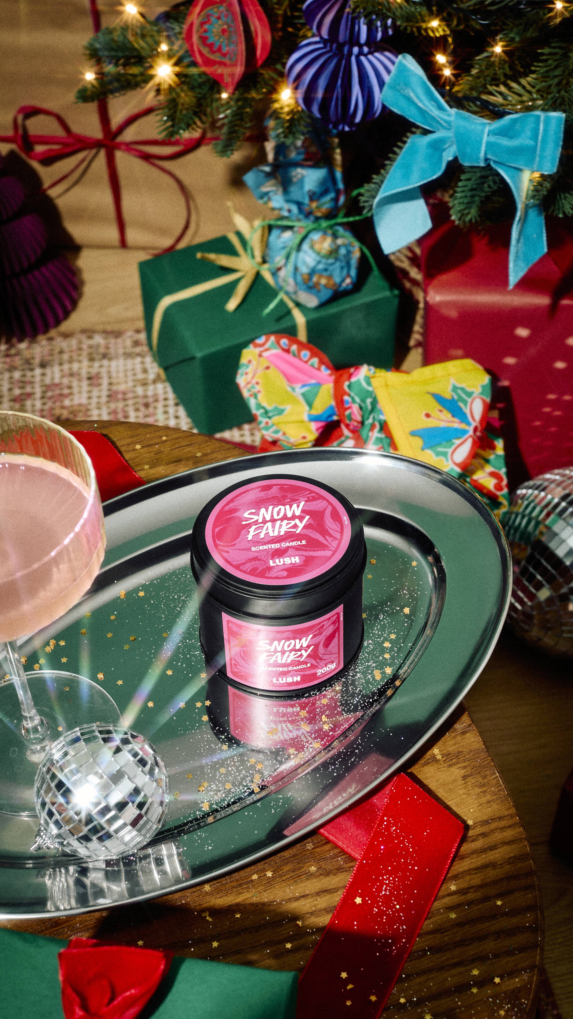 A black tin of Lush Snow Fairy scented candle with a pink label sits on a silver tray with gold glitter, alongside a cocktail glass and mini disco ball, set against a festive backdrop of wrapped gifts and a Christmas tree.