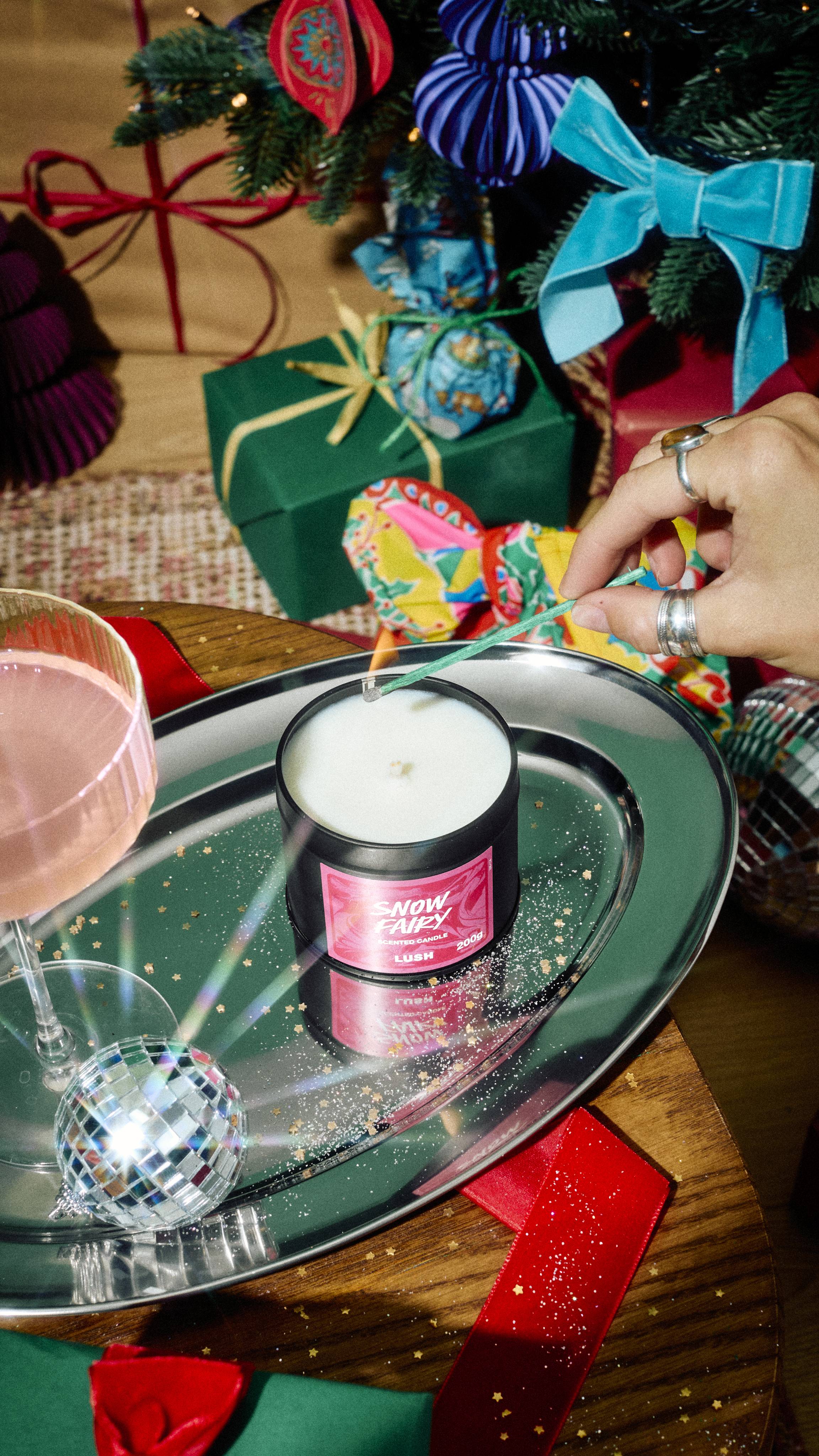 A hand uses a long match to light an open tin of Lush Snow Fairy scented candle on a silver tray with gold glitter, surrounded by a cocktail glass, a mini disco ball, and festive wrapped gifts in the background.