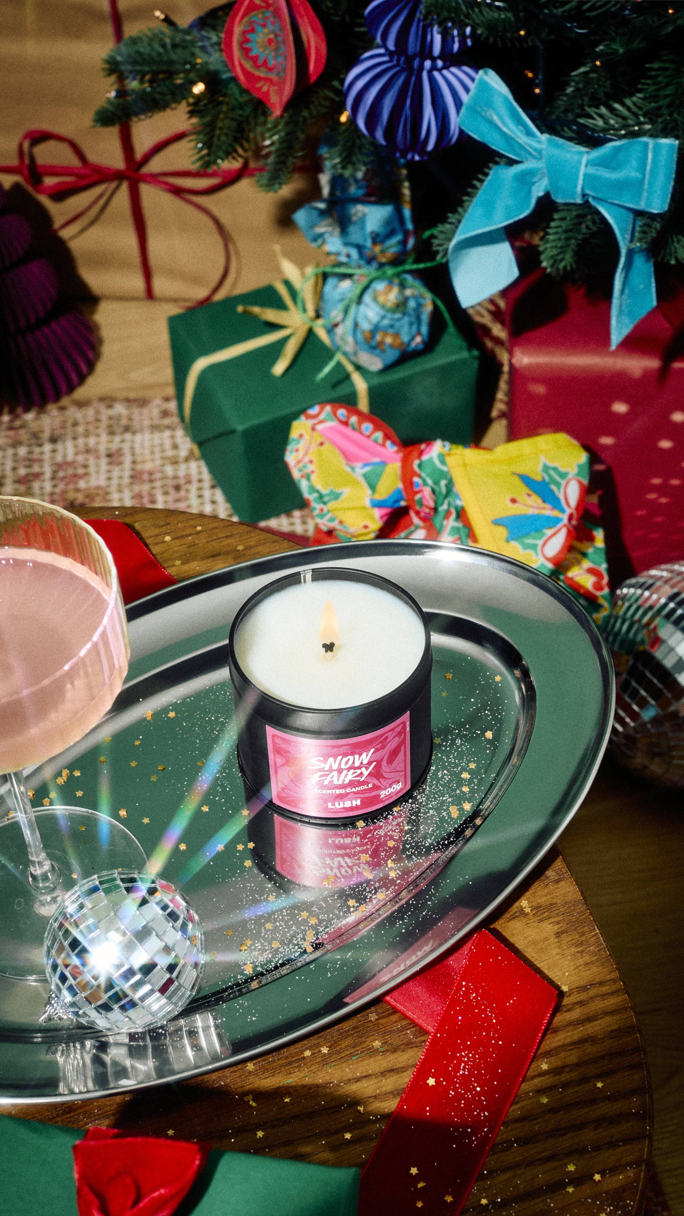 A lit tin of Lush Snow Fairy scented candle sits burning on a silver tray scattered with gold glitter, beside a cocktail glass and mini disco ball, set against a festive background of wrapped presents.