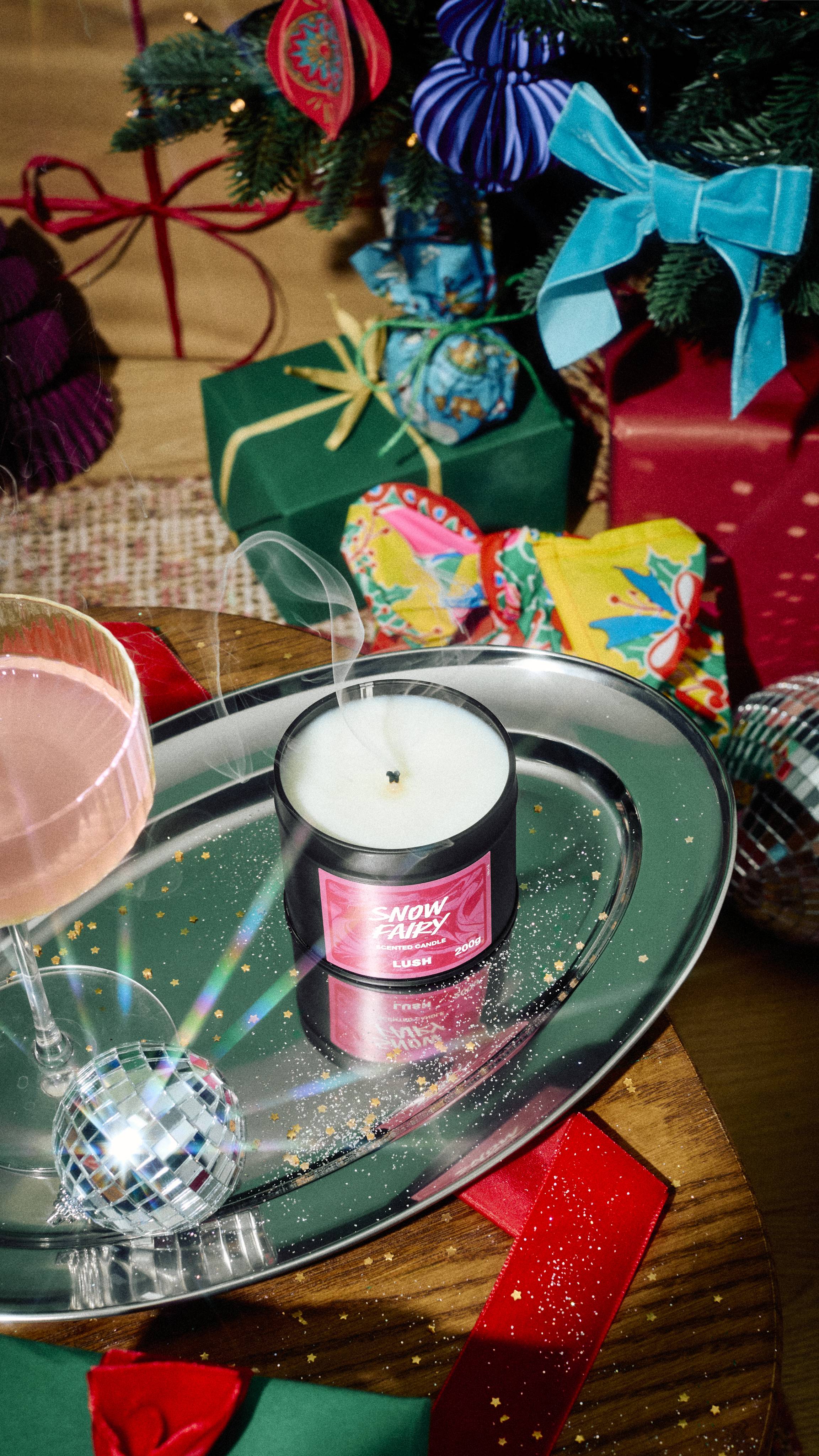 Smoke rises from a freshly extinguished tin of Lush Snow Fairy scented candle on a silver tray with gold glitter, alongside a cocktail glass and mini disco ball, set against a festive backdrop of wrapped gifts.