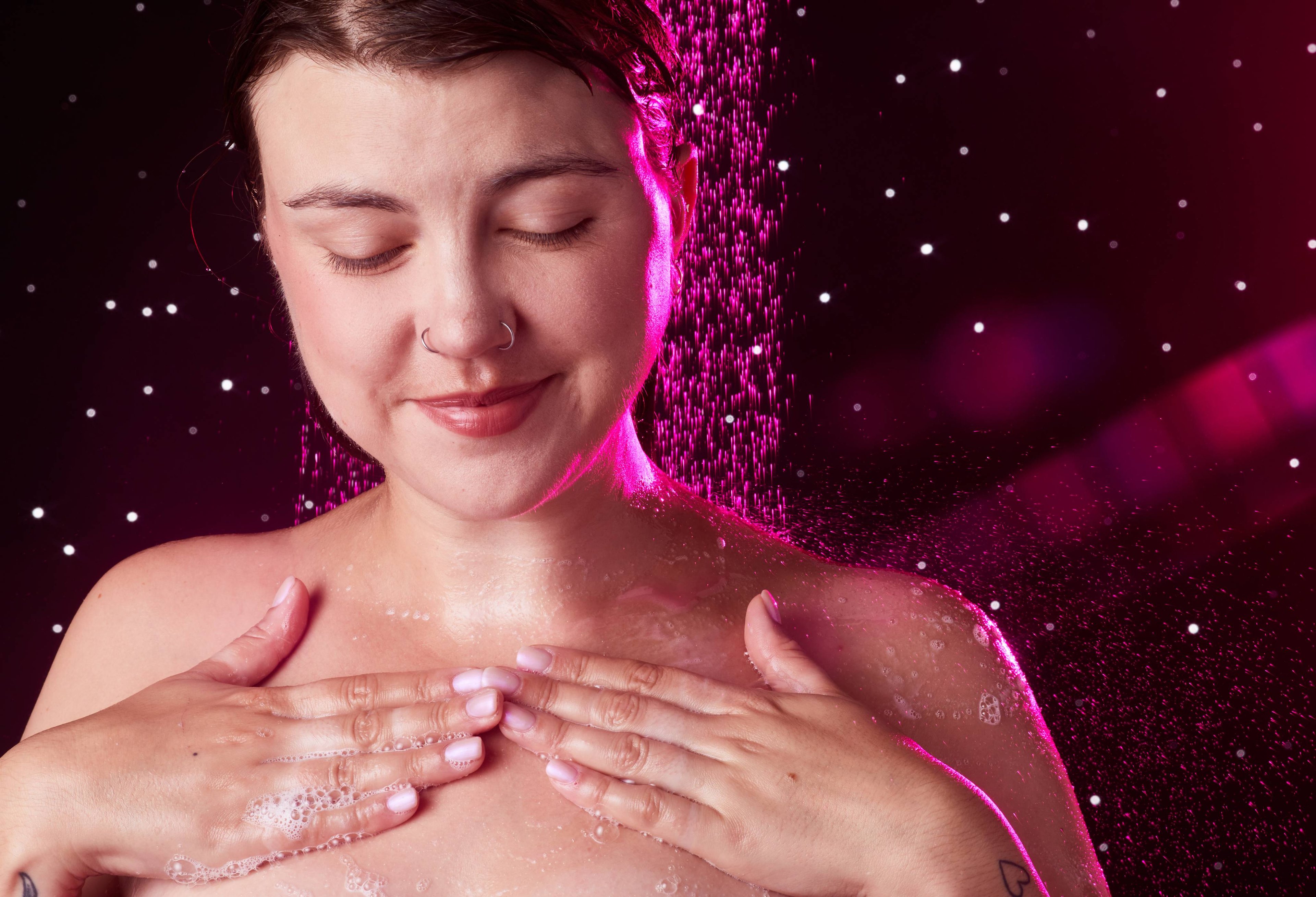 Image shows the model on a twinkling background under running shower water as they later up their chest with both hands. 