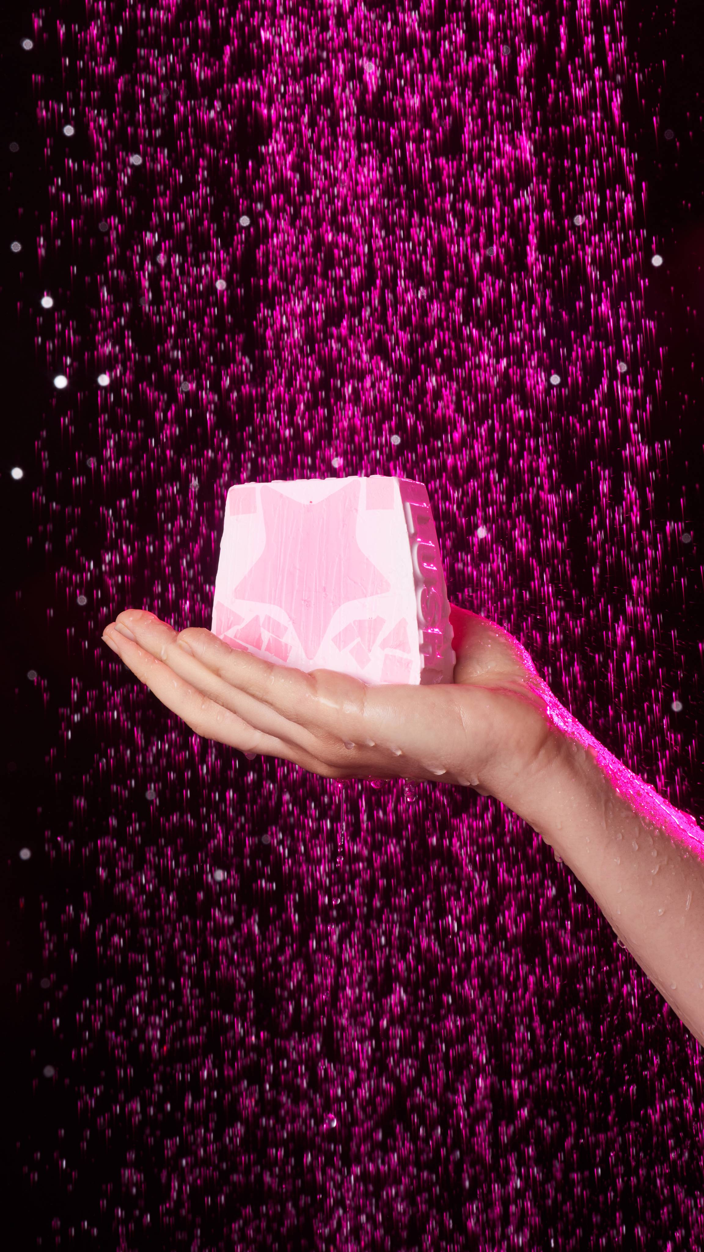 A close-up of a single hand holding the Snow Fairy soap as shower water falls, tinted by pink lighting.