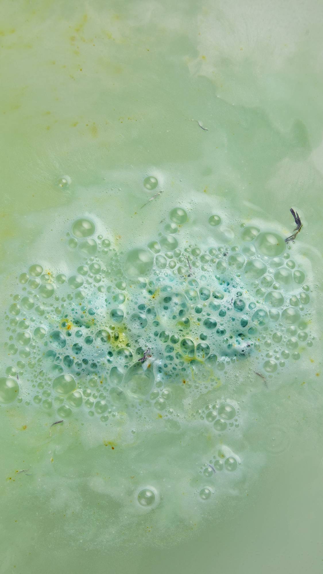 The image shows the Lush Snow Queen bath bomb has almost fully dissolved in delicate bath water. There is a slowly dissolving clump of soft flue with speckles of yellow and dried flowers bursting from the centre. 