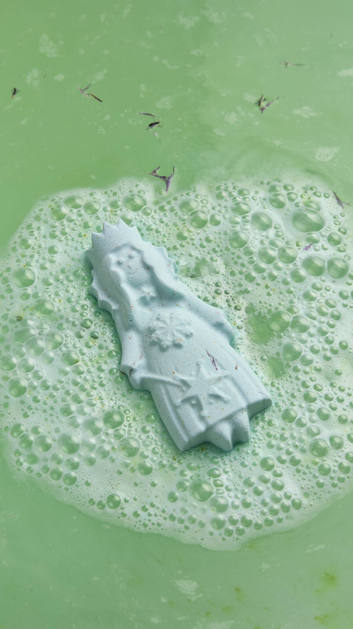 An angled, aerial shot of the Lush Snow Queen bath bomb sitting on the surface of the water as it begins to dissolve. There is a light foam coming from the bath bomb and delicate, dried flowers have emerged from the product.