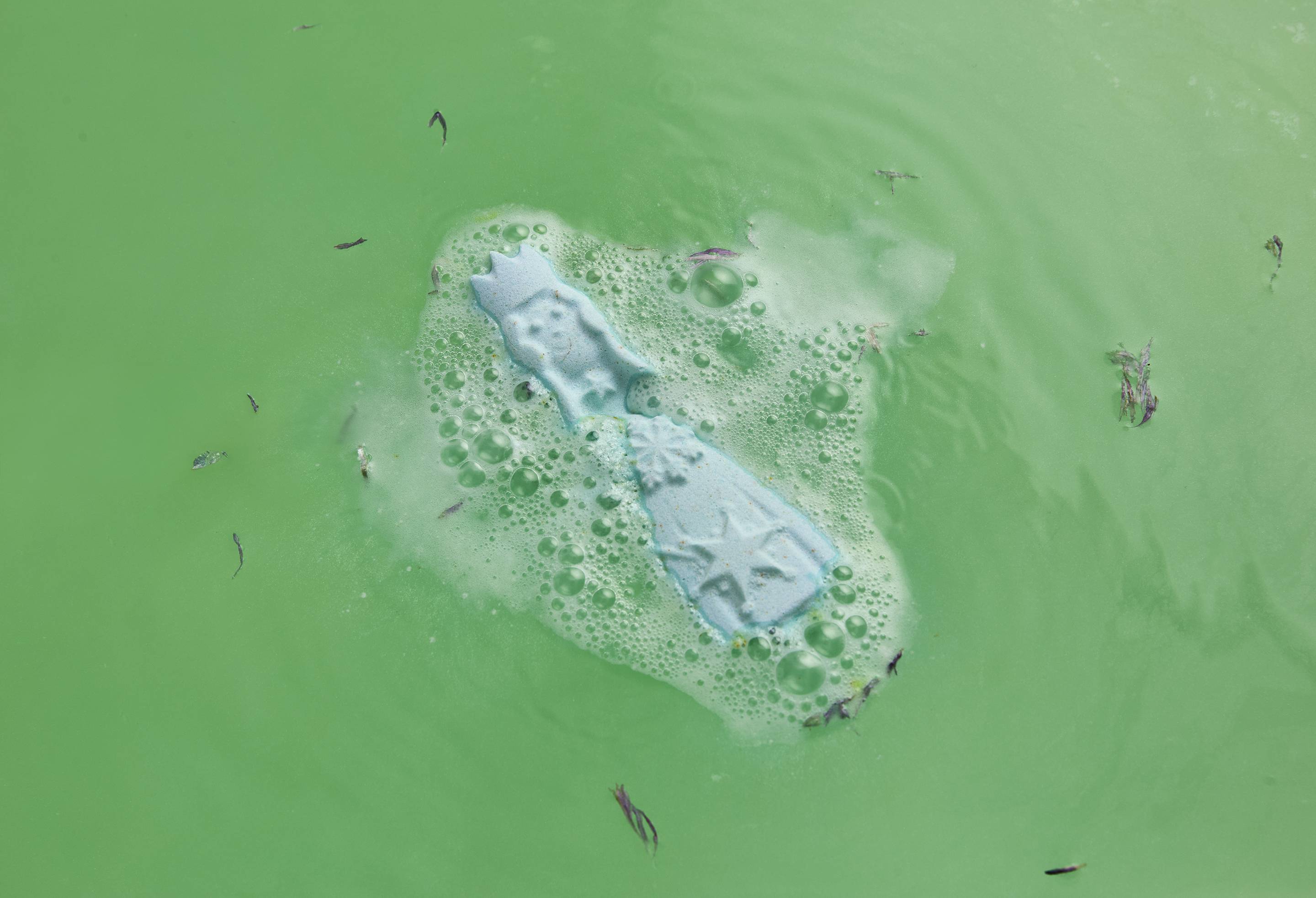 An angled, aerial shot of the Lush Snow Queen bath bomb sitting on the surface of the water as it begins to dissolve. There is a light foam coming from the bath bomb and delicate, dried flowers have emerged from the product.
