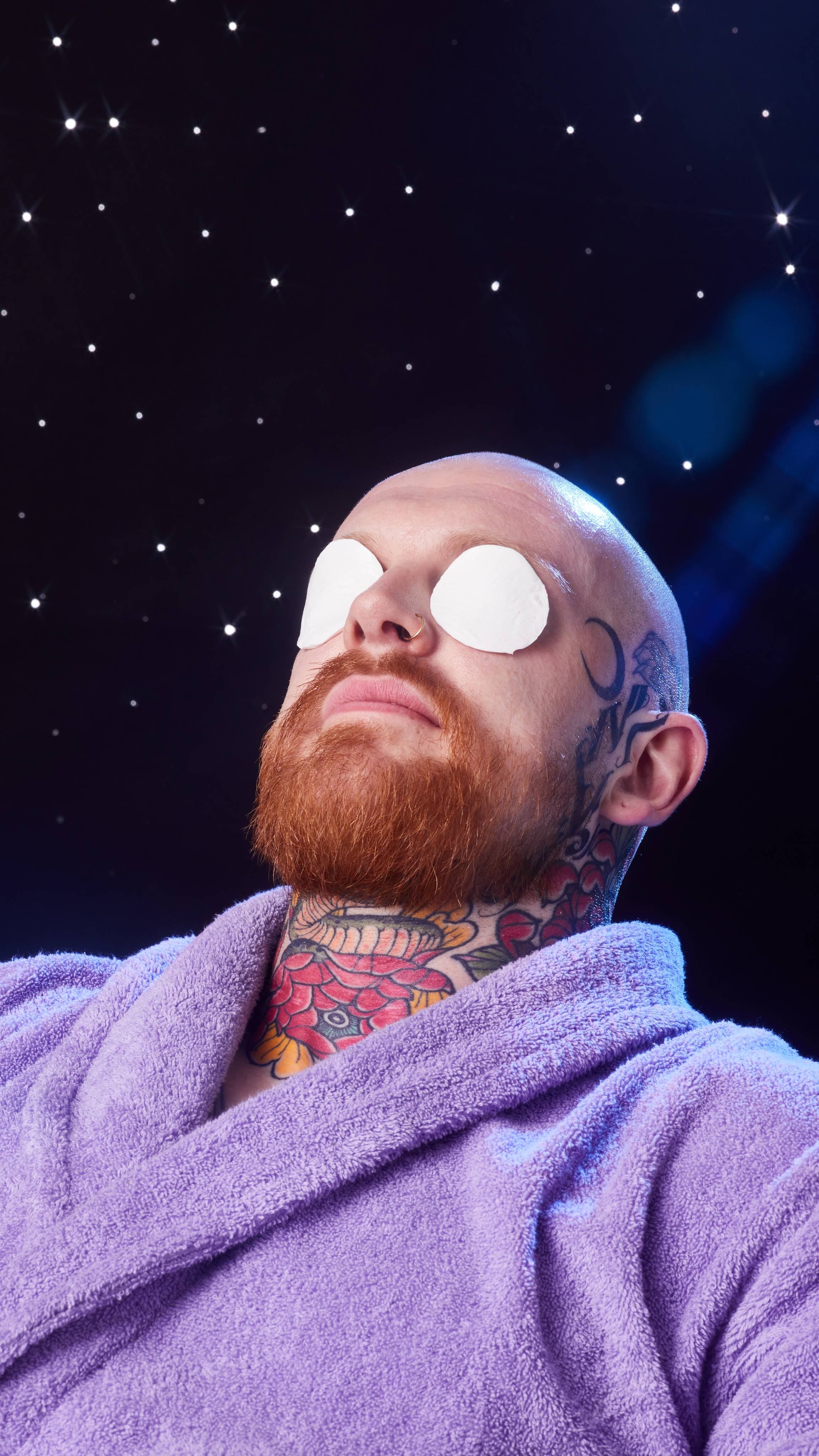 Model sits in a purple robe on a starry backdrop with the white Snowball eye pads laid over their eyes. 