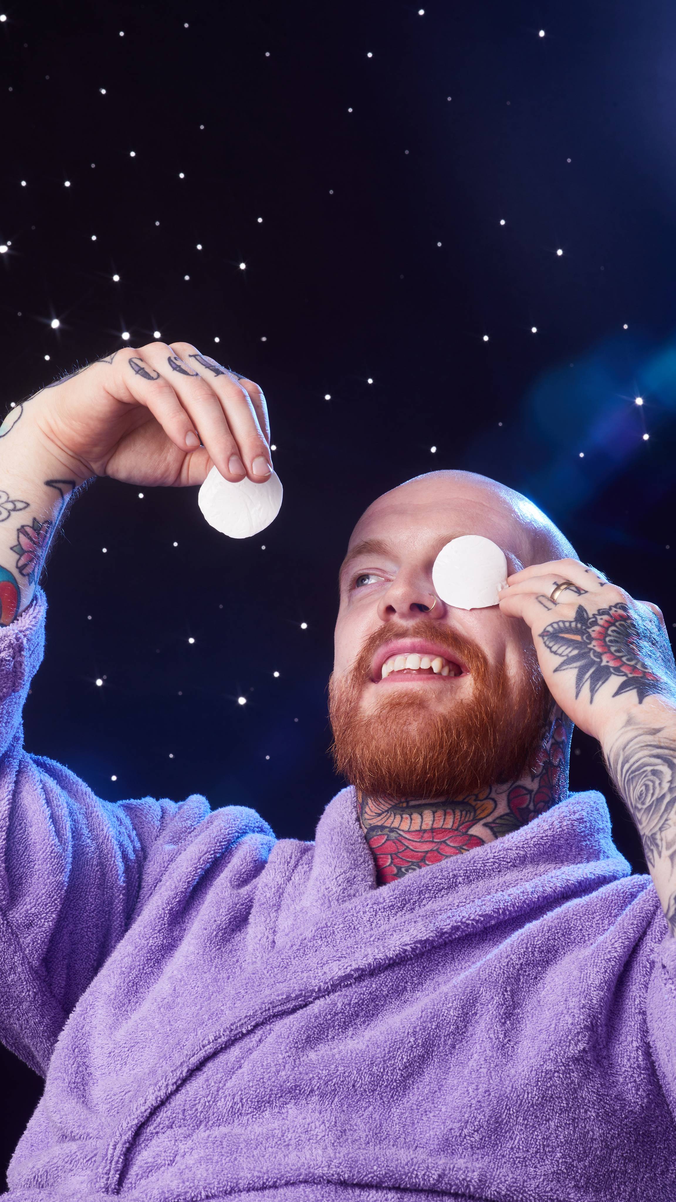 Model is in a purple robe on a starry backdrop and is smiling as they hold the Snowball eye pads in front of their eyes. 