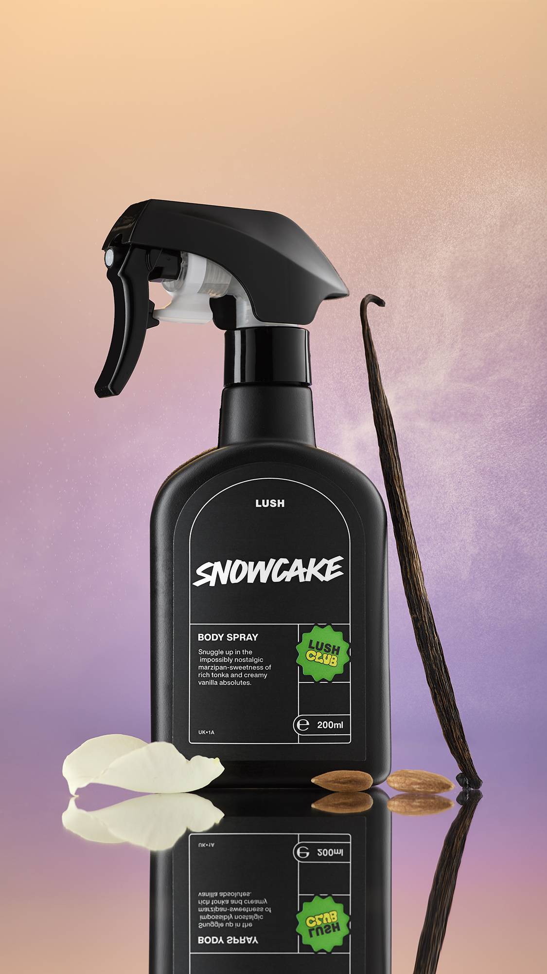 
A black spray bottle of Snowcake Body Spray stands on a reflective surface against a soft orange and lavender gradient background. It is flanked by a vanilla bean, an almond, and a creamy white petal.