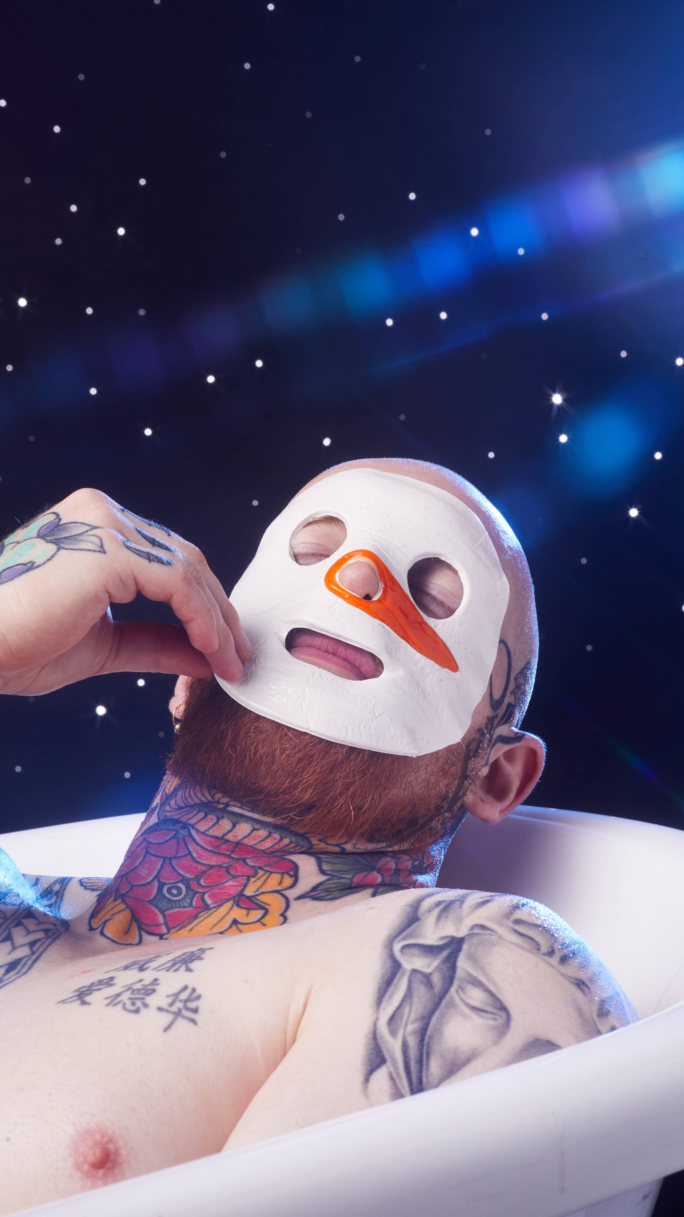 The model lays in a bathtub on a starry background positioning the Snowman sheet mask gently on their face.
