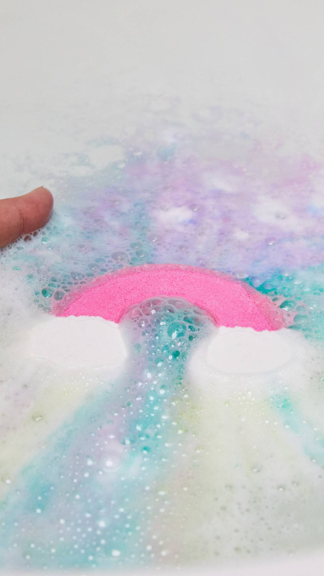A close-up of the Somewhere bath bomb being placed into the bath water as it immediately ripples out pastel blue, pink and purple.