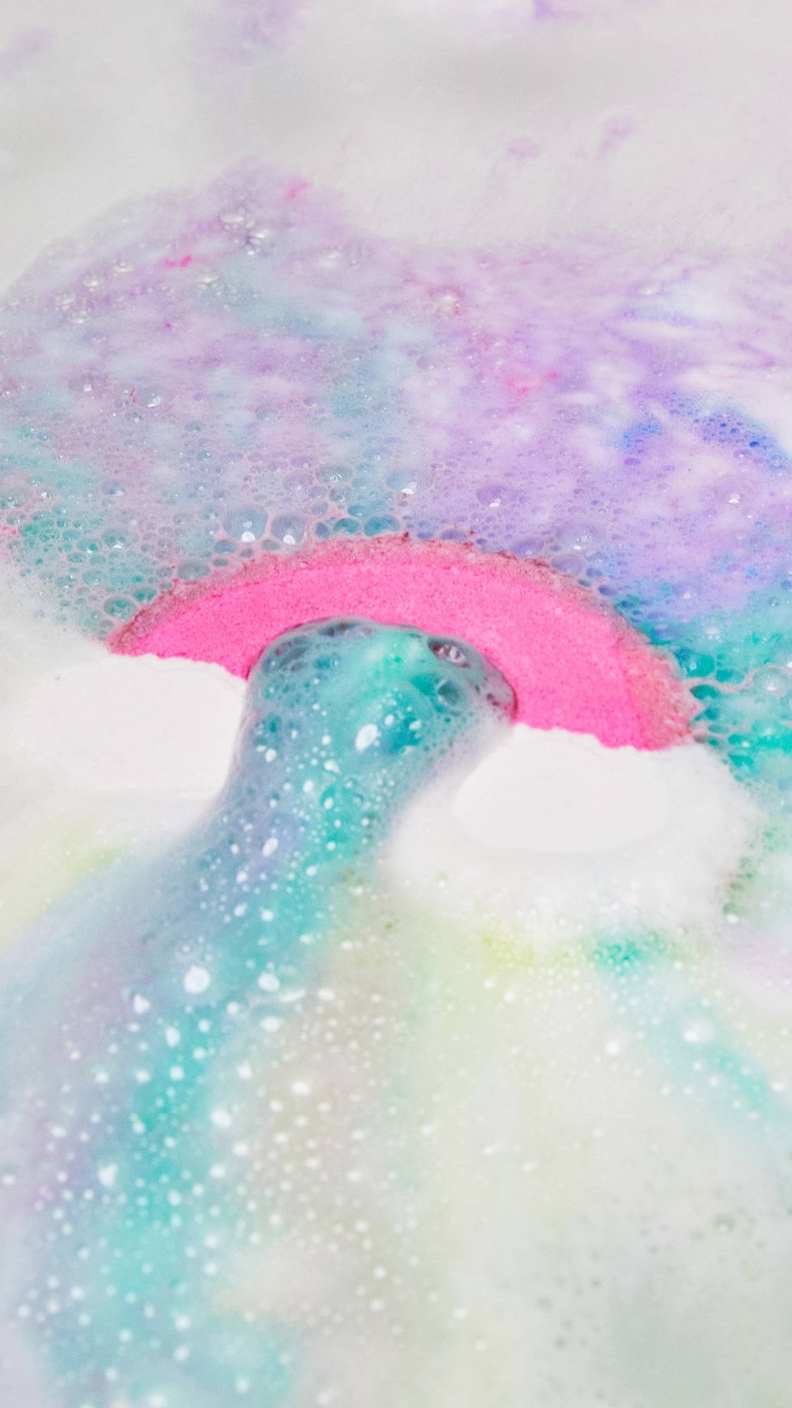 A close-up of the Somewhere bath bomb being placed into the bath water as it immediately ripples out pastel blue, pink and purple.