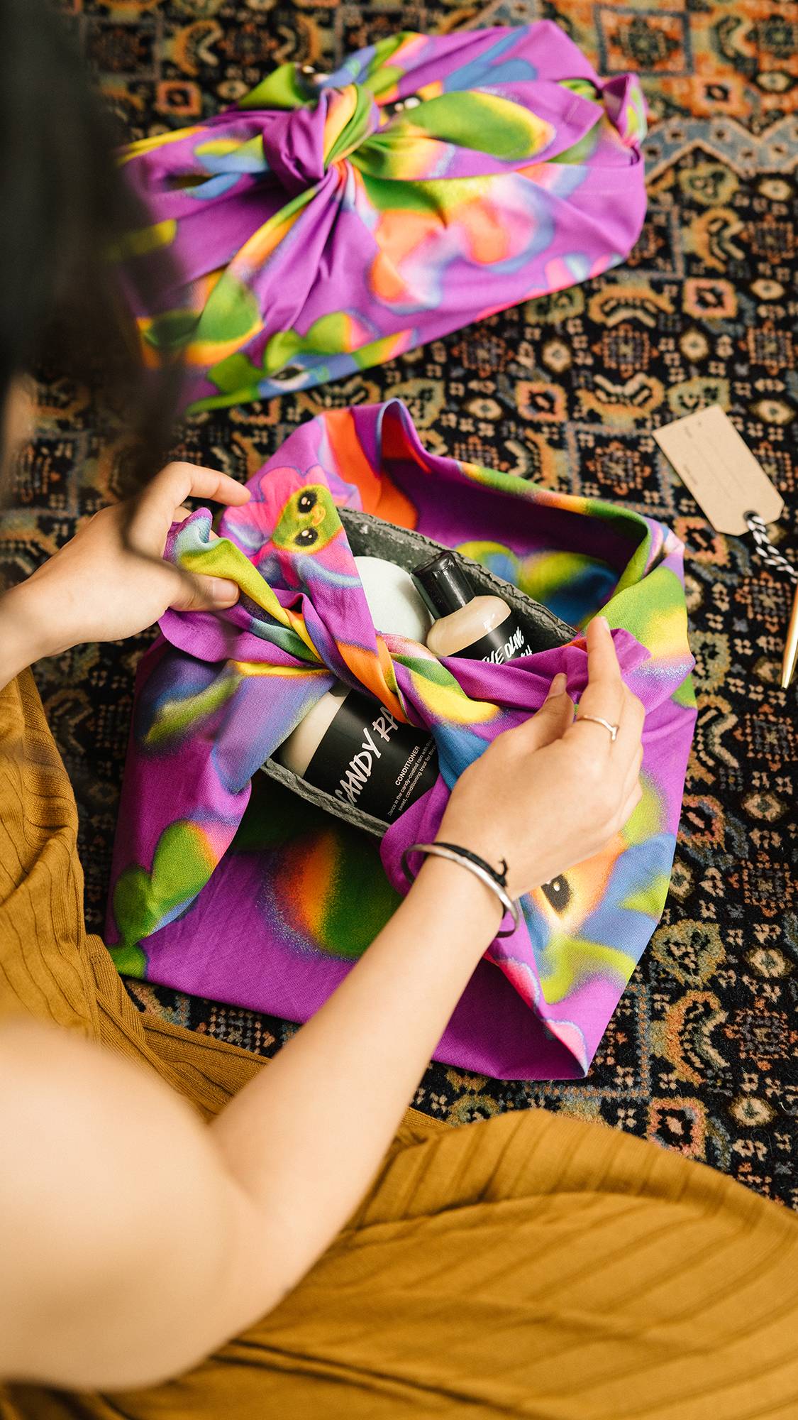 Image shows the model sat on the floor midway through wrapping a LUSH gift with the Soopa Frens colourful knot wrap.