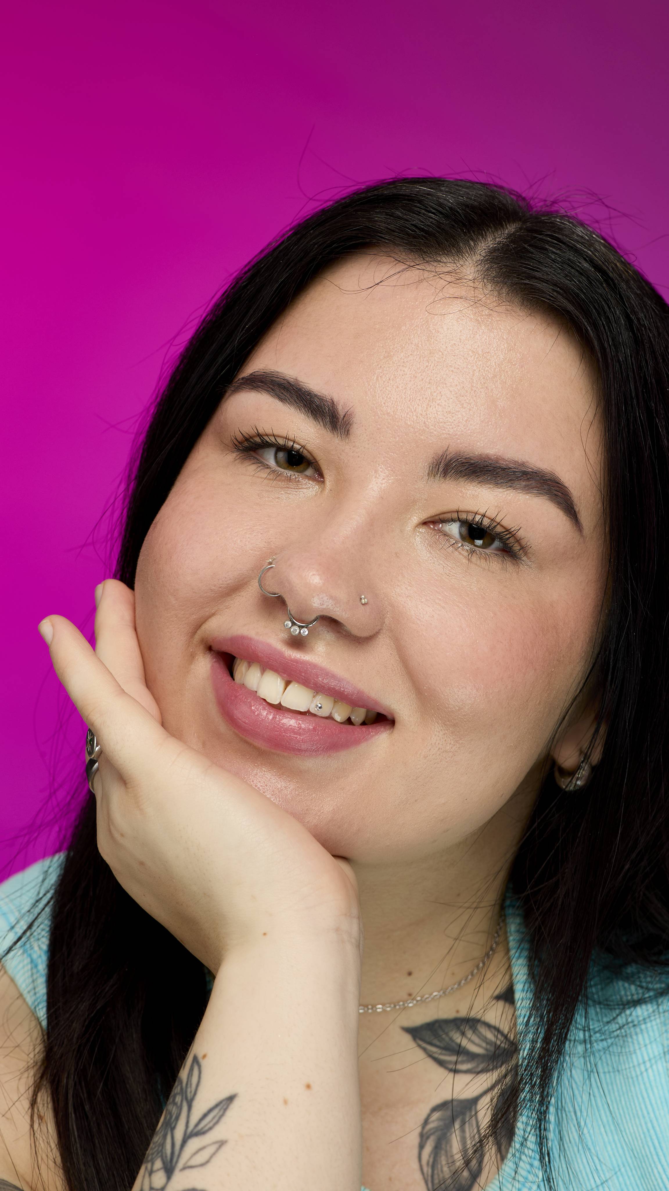 A smiling person with long, dark hair, wearing a light blue ribbed top, is shown from the chest up. They are on a vibrant pink background, showing off the Sparkler lipstick with a subtle glittering shimmer as they rest on their hand.