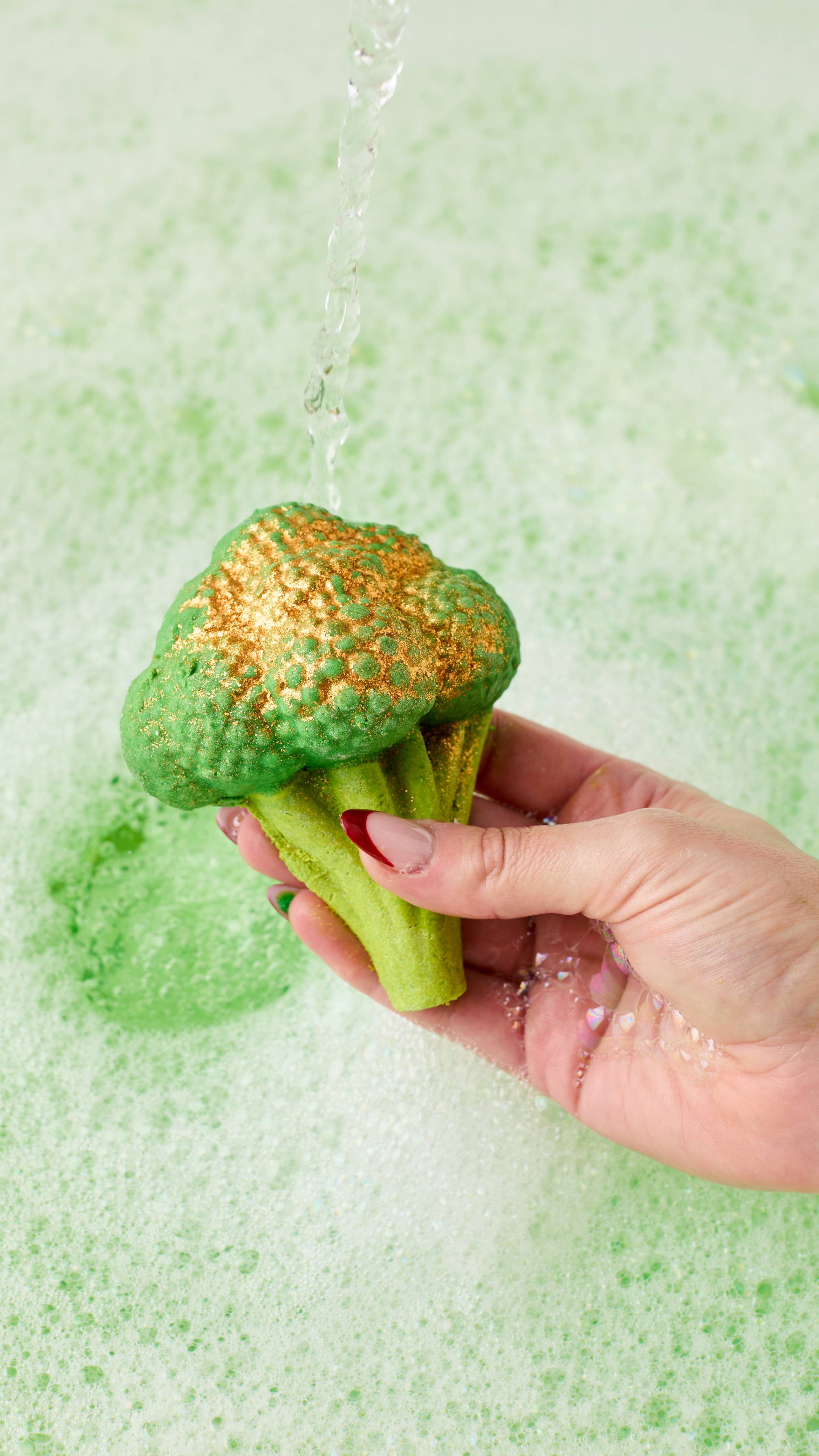 The image shows a hand with long nails holding the vibrant green Sparkly Broccoli bubble bar under a stream of water. Delicate green water sits below, covered in a blanket of thick bubbles. 